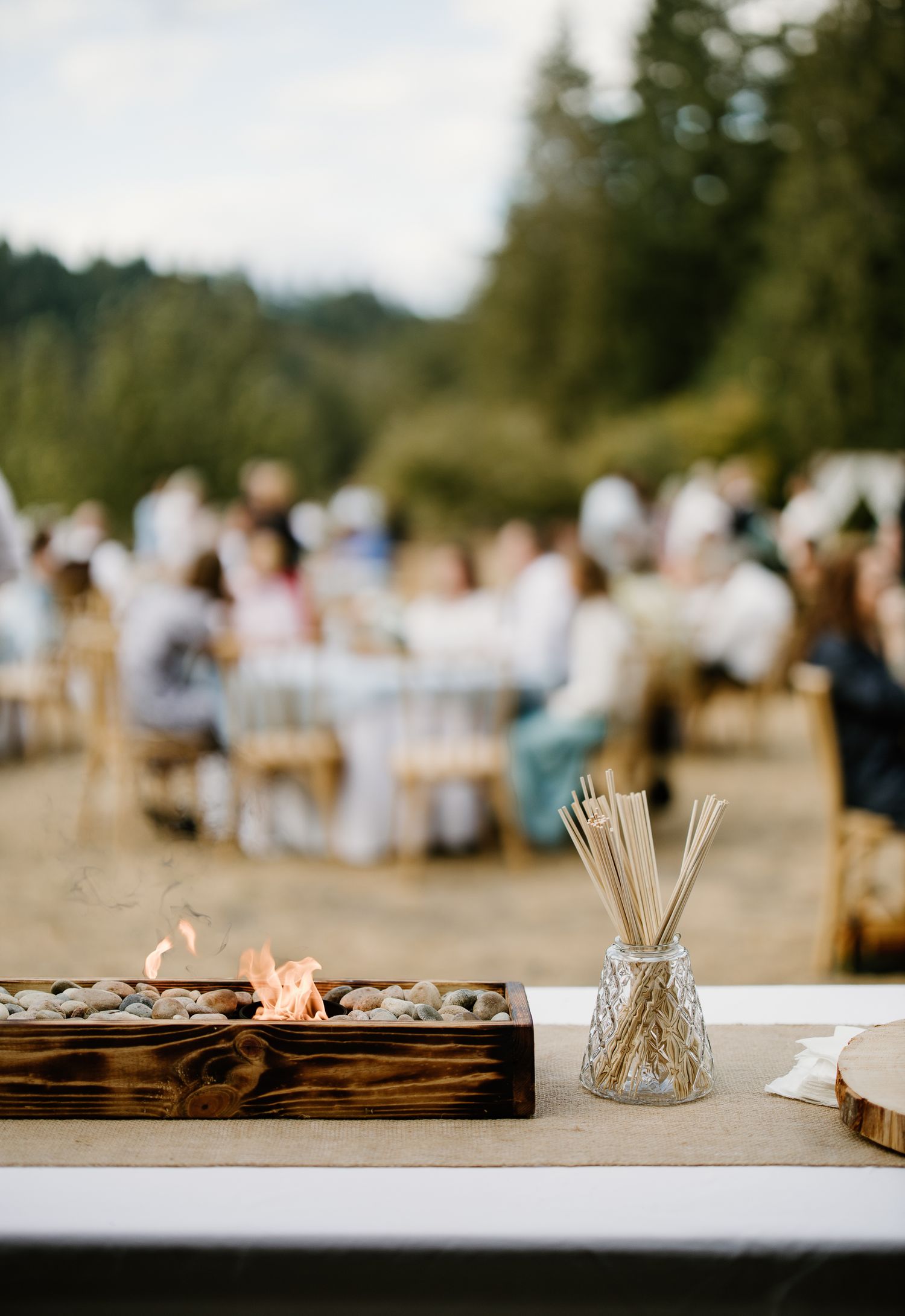 Outdoor wedding reception with a wooden fire pit centerpiece and skewers in a glass vase, guests dining al fresco.