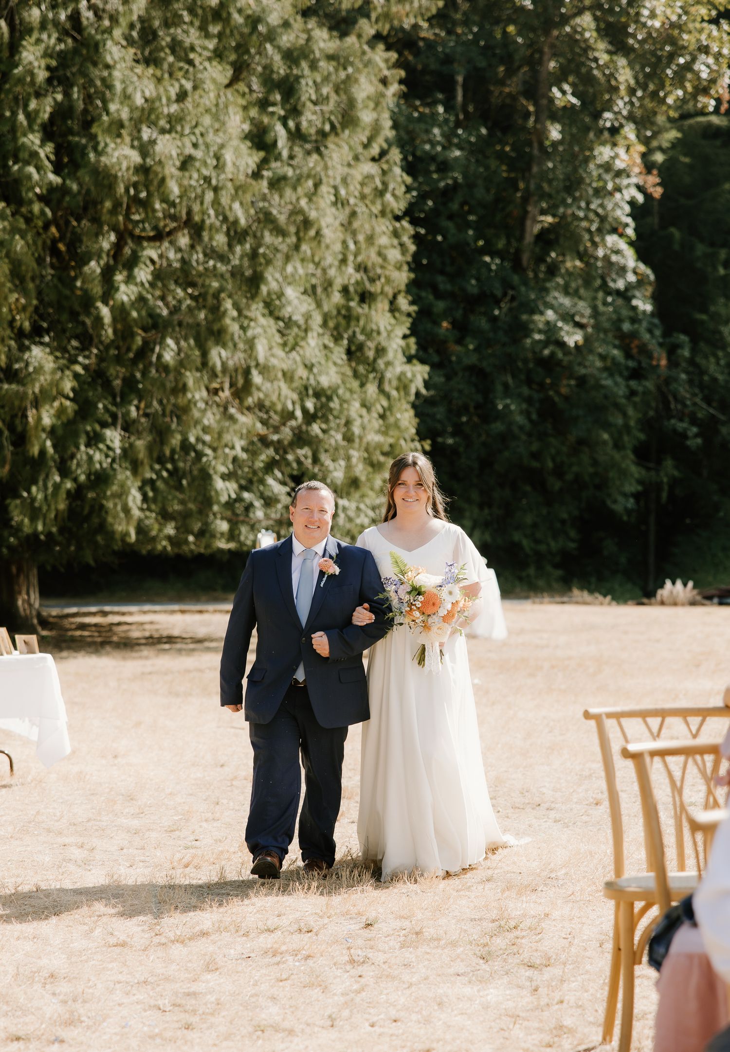 Bride walks down outdoor aisle with escort, holding colorful bouquet, surrounded by lush green trees.