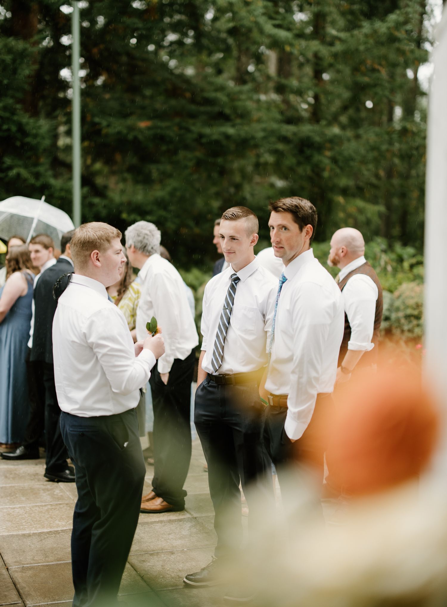 Groomsmen in white shirts and ties socializing outdoors at a wedding reception surrounded by tall evergreen trees.