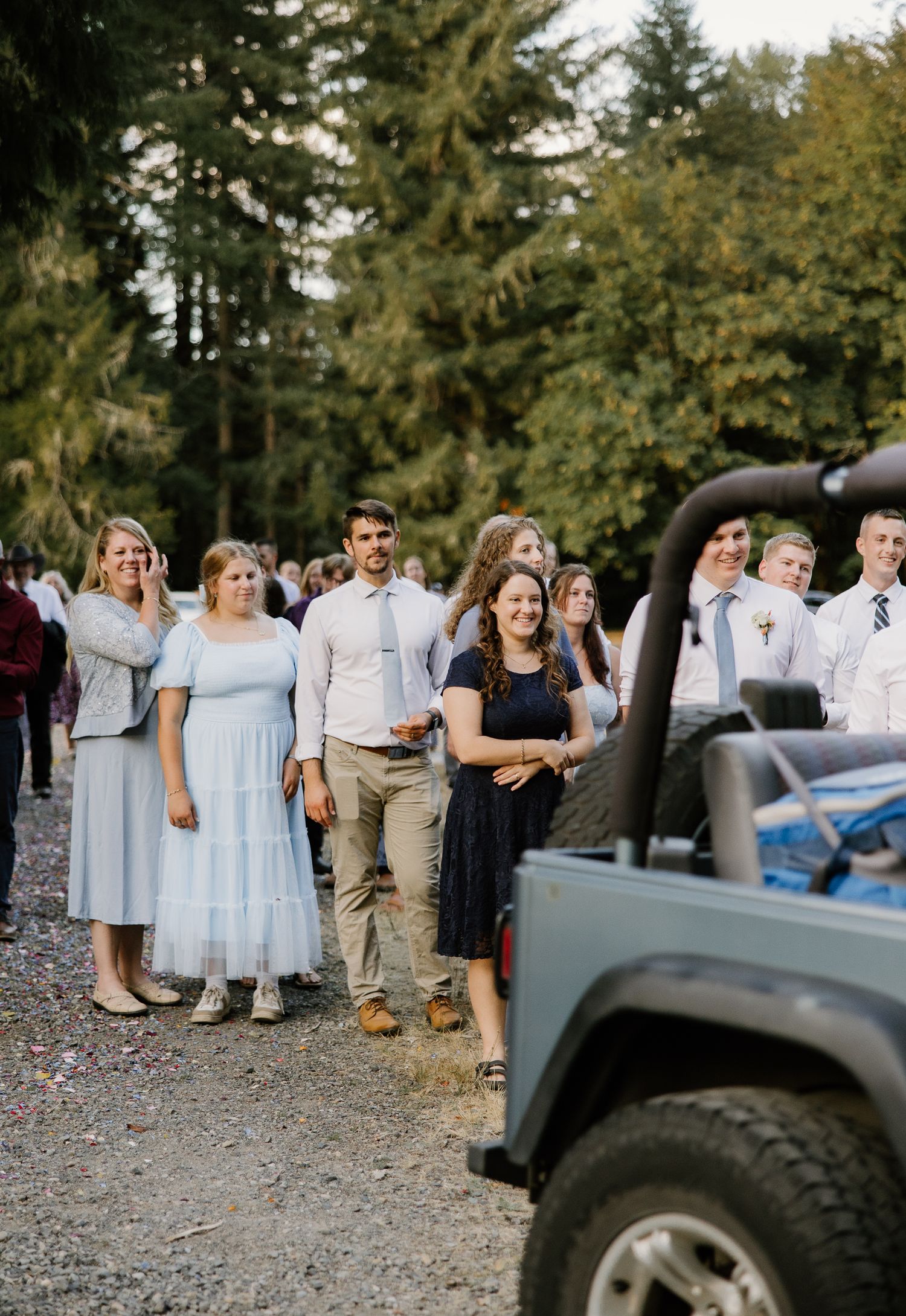 Wedding guests gather on gravel path near blue Jeep after outdoor ceremony in forest setting.
