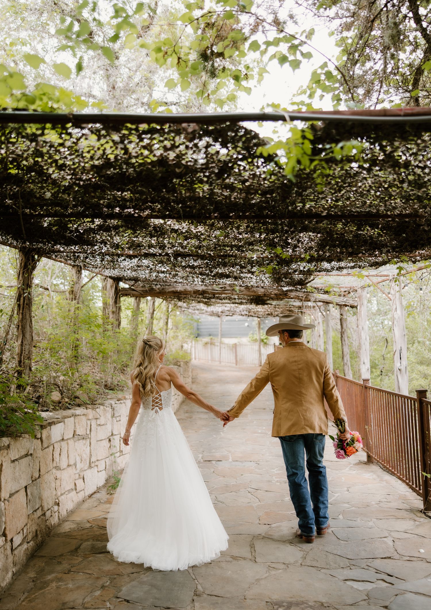 Bride and groom in cowboy hat walk hand-in-hand under vine-covered pergola on stone path at rustic wedding.