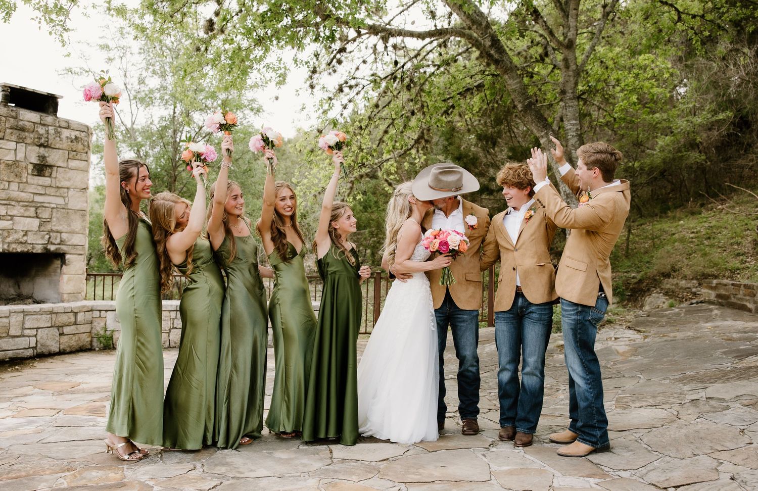 Bridal party celebrating outdoors with bridesmaids in green dresses and groomsmen in tan blazers and cowboy hats.