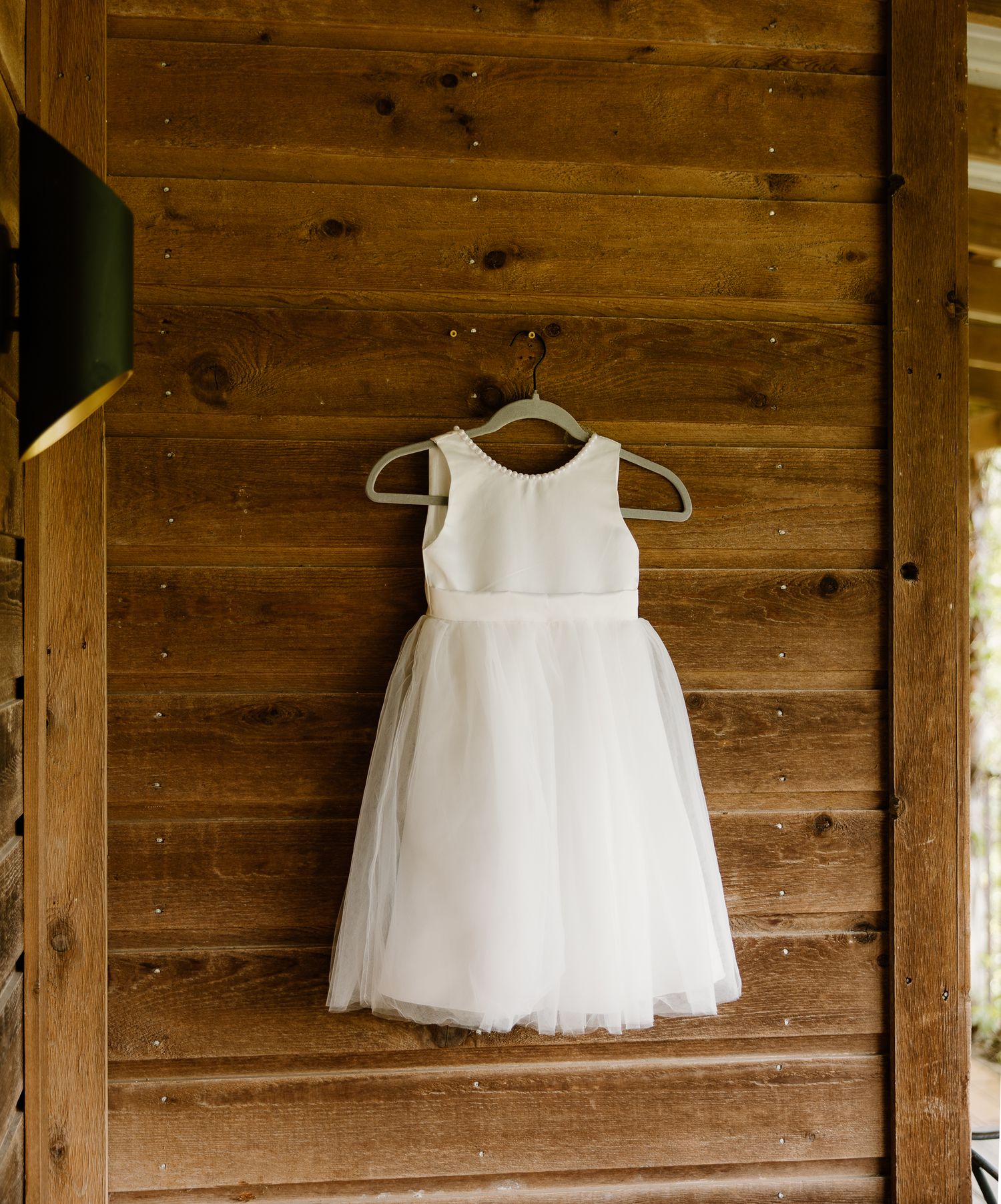 White flower girl dress hanging on wooden wall at rustic wedding venue.
