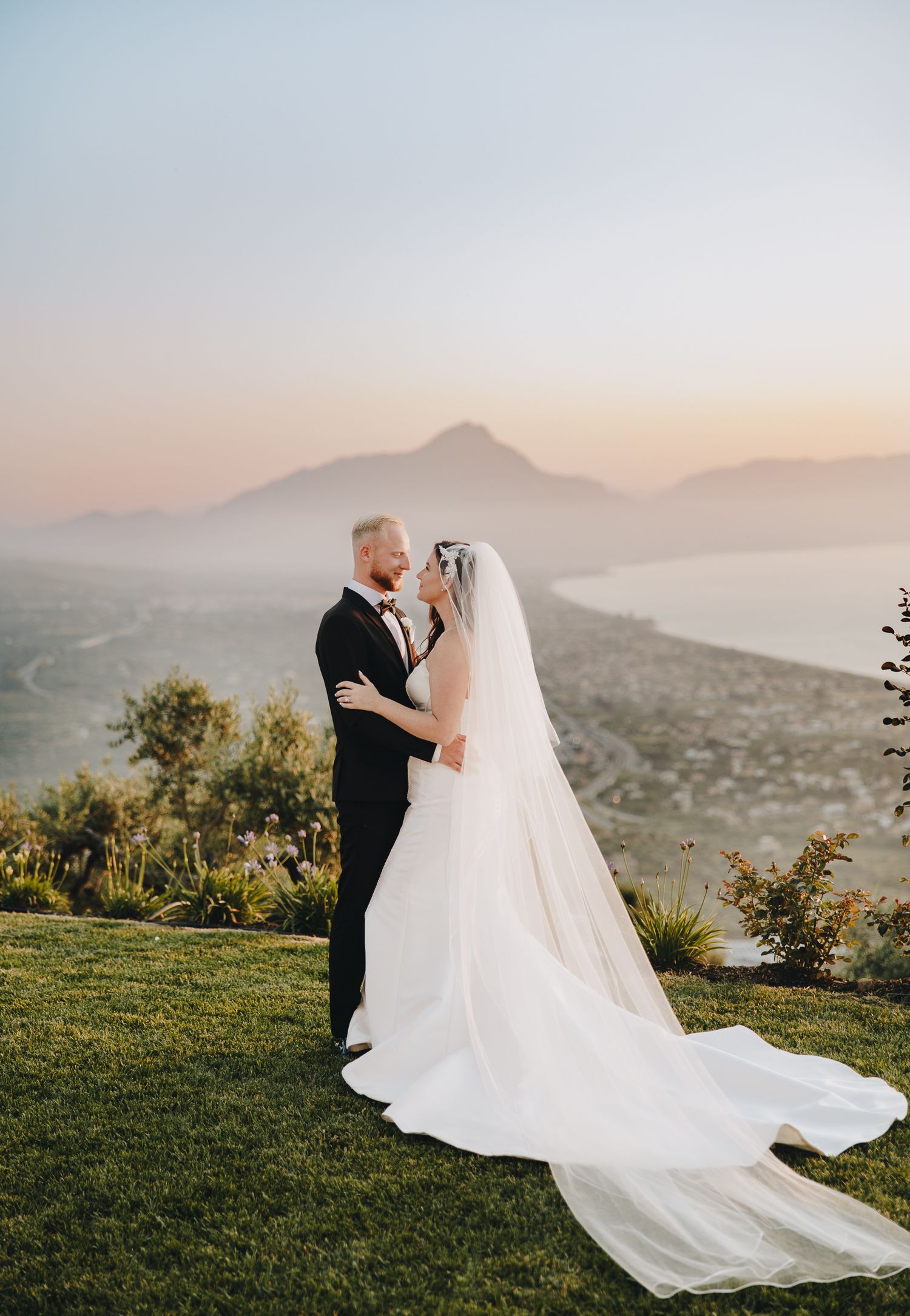 Bride and groom embrace at sunset with breathtaking mountain lake panoramic view behind them.