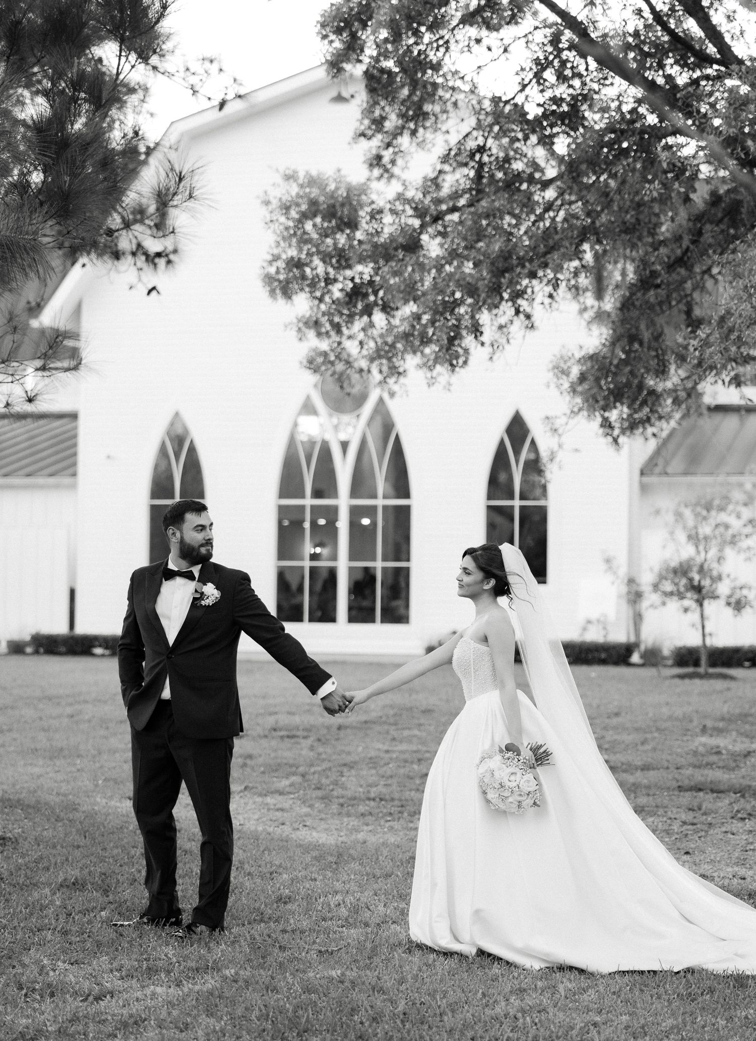 Black and white portrait of bride and groom holding hands before a white chapel.