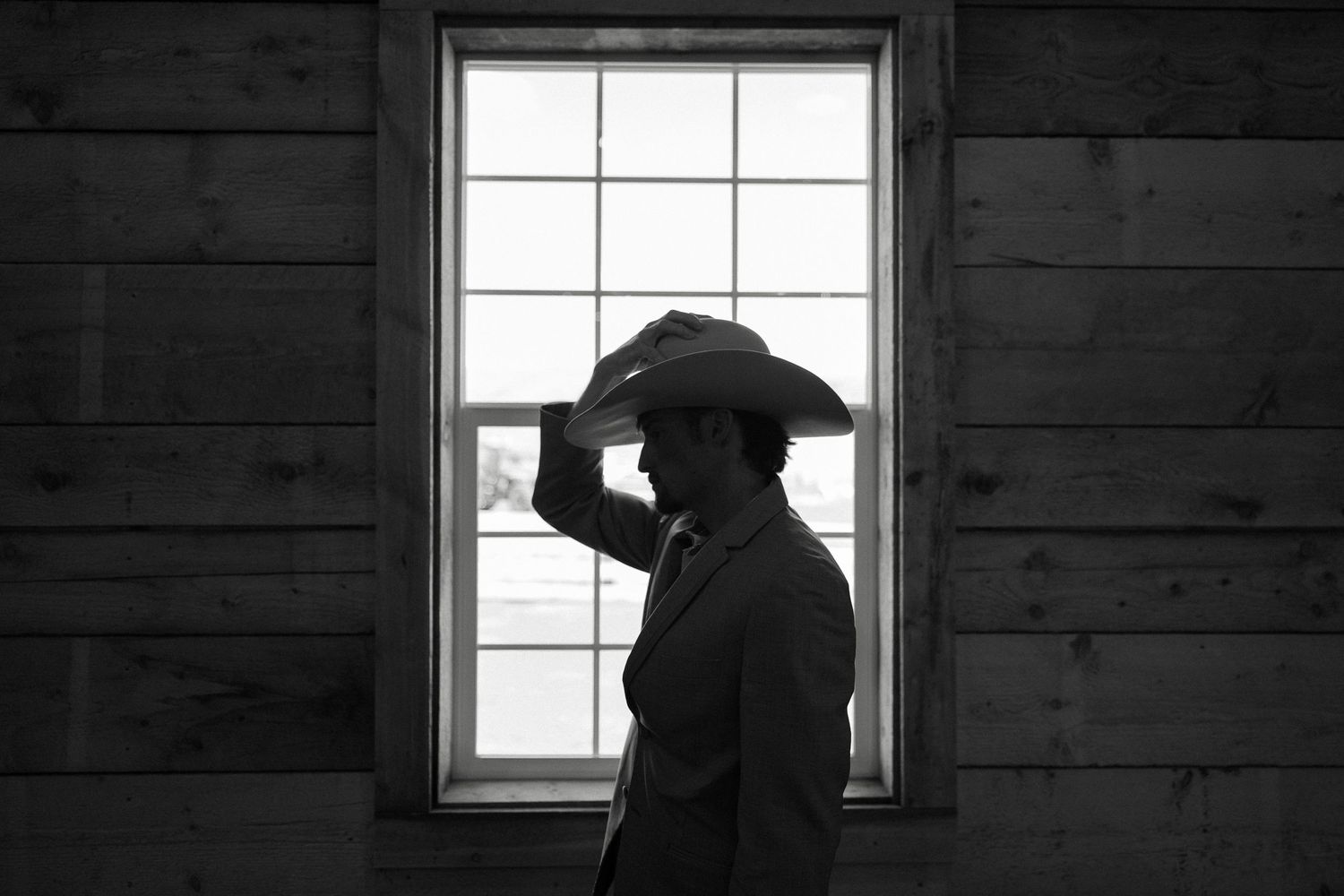 Black and white silhouette of a man in a cowboy hat standing before a window in a rustic wooden barn.