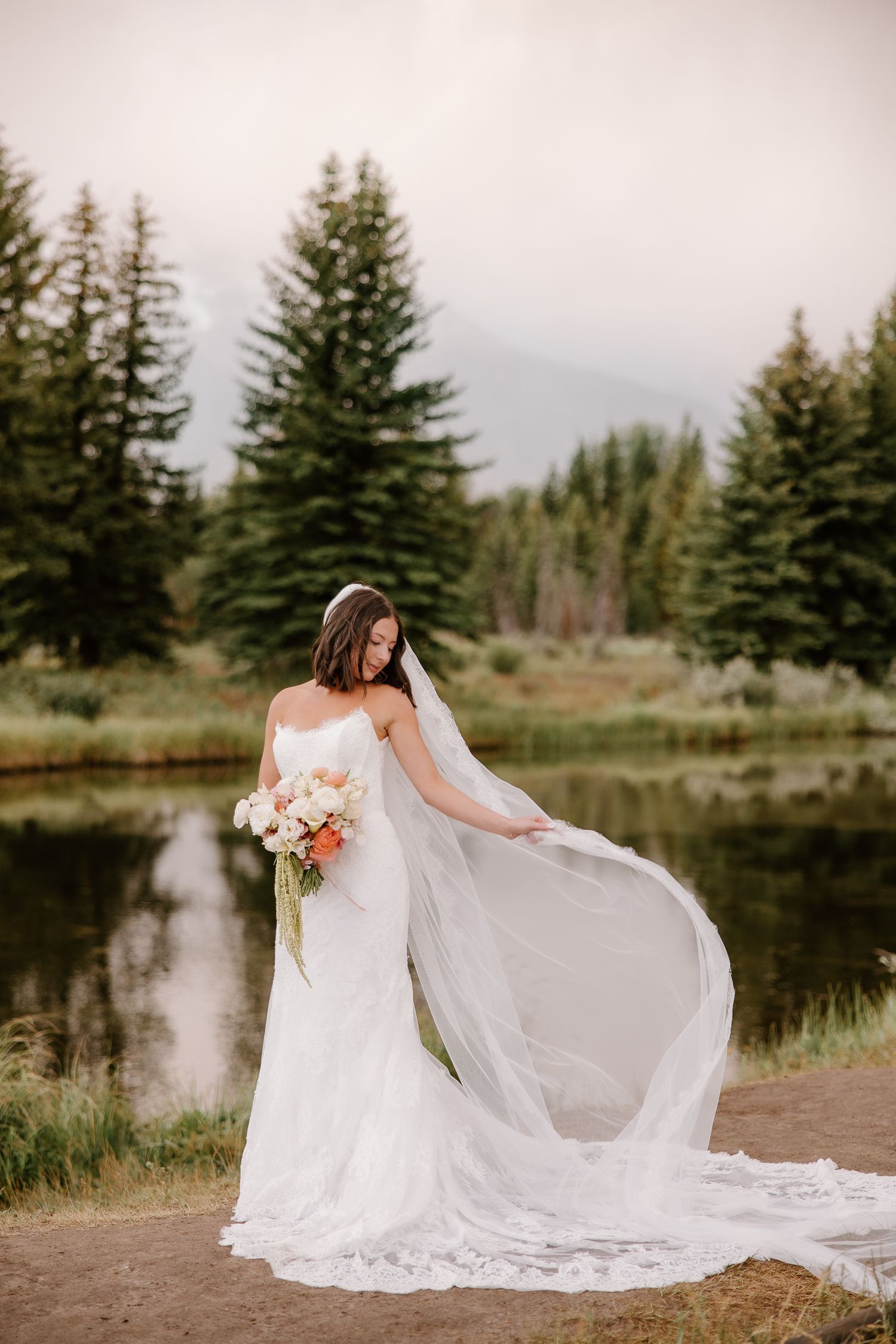 Bride in white lace gown holding floral bouquet, veil flowing by mountain lake with evergreen trees backdrop.