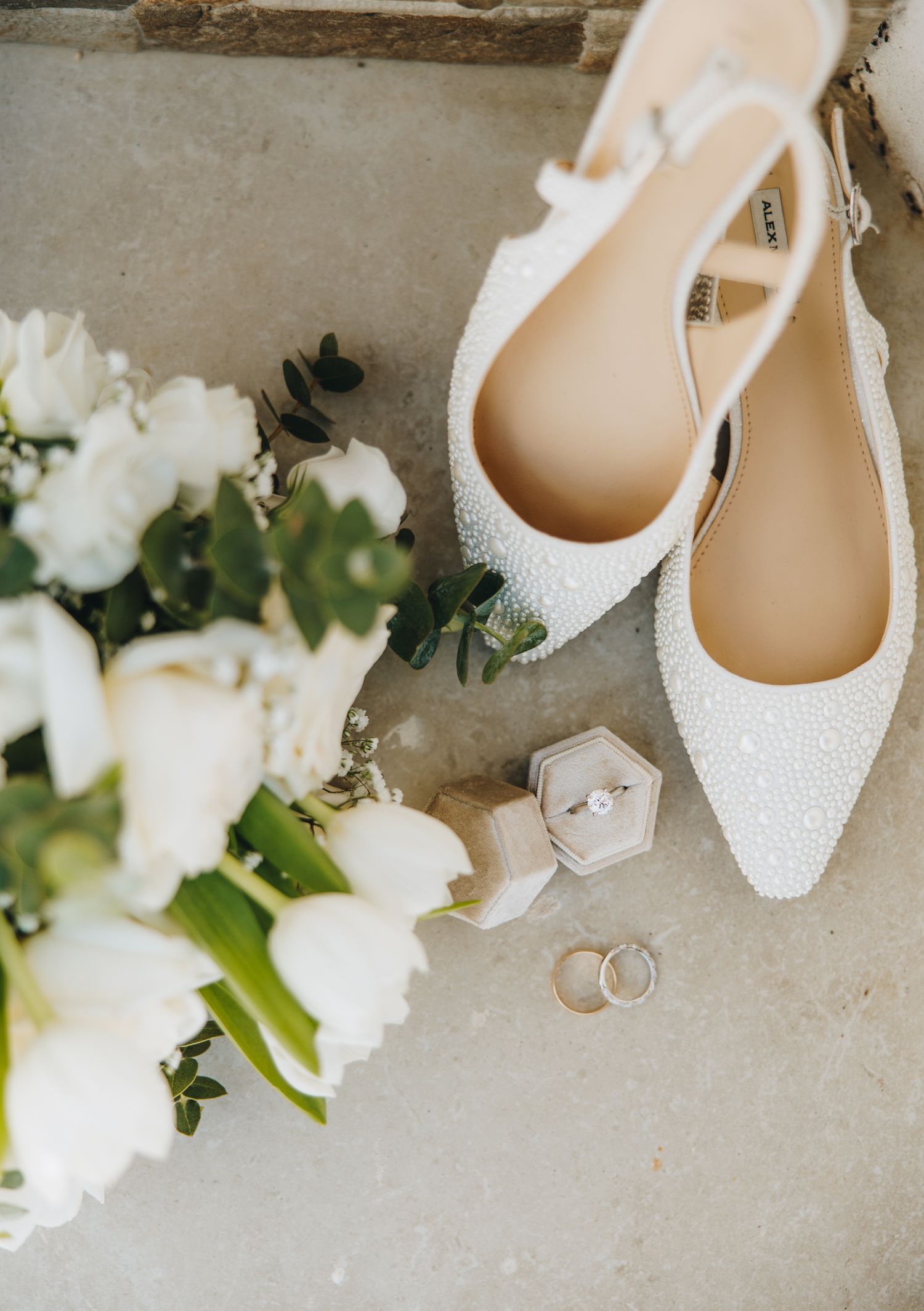 White beaded bridal heels, wedding rings, and white floral bouquet arranged on stone surface.
