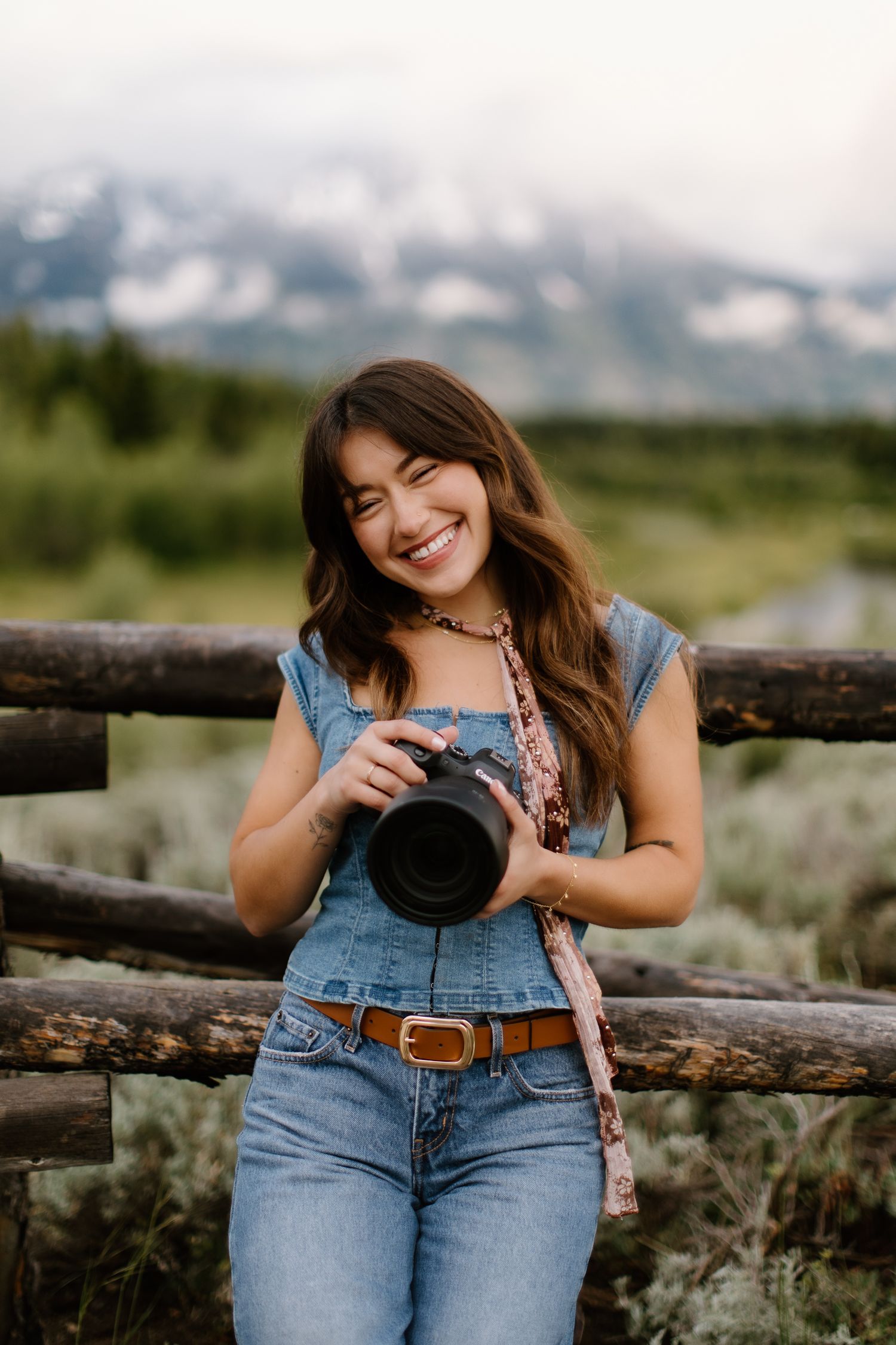 Smiling young woman in denim overalls holding a camera, leaning on a rustic wooden fence with mountains in the background.