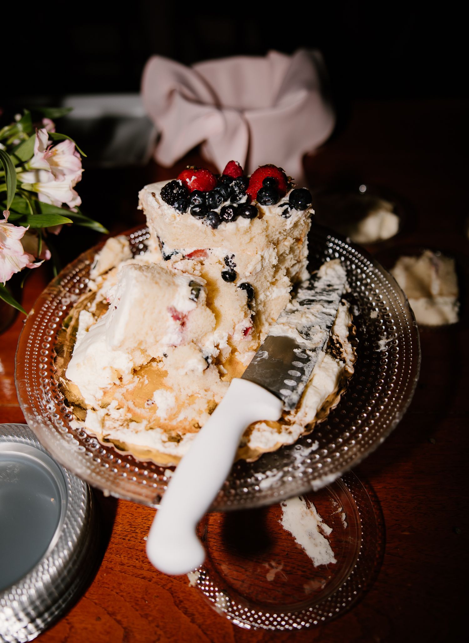 A partially sliced wedding cake topped with fresh berries sits on a glass cake stand on a wooden table.