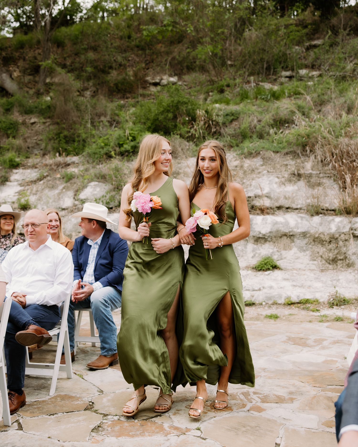 Two bridesmaids in olive green satin dresses walk down the aisle holding colorful bouquets at an outdoor wedding.