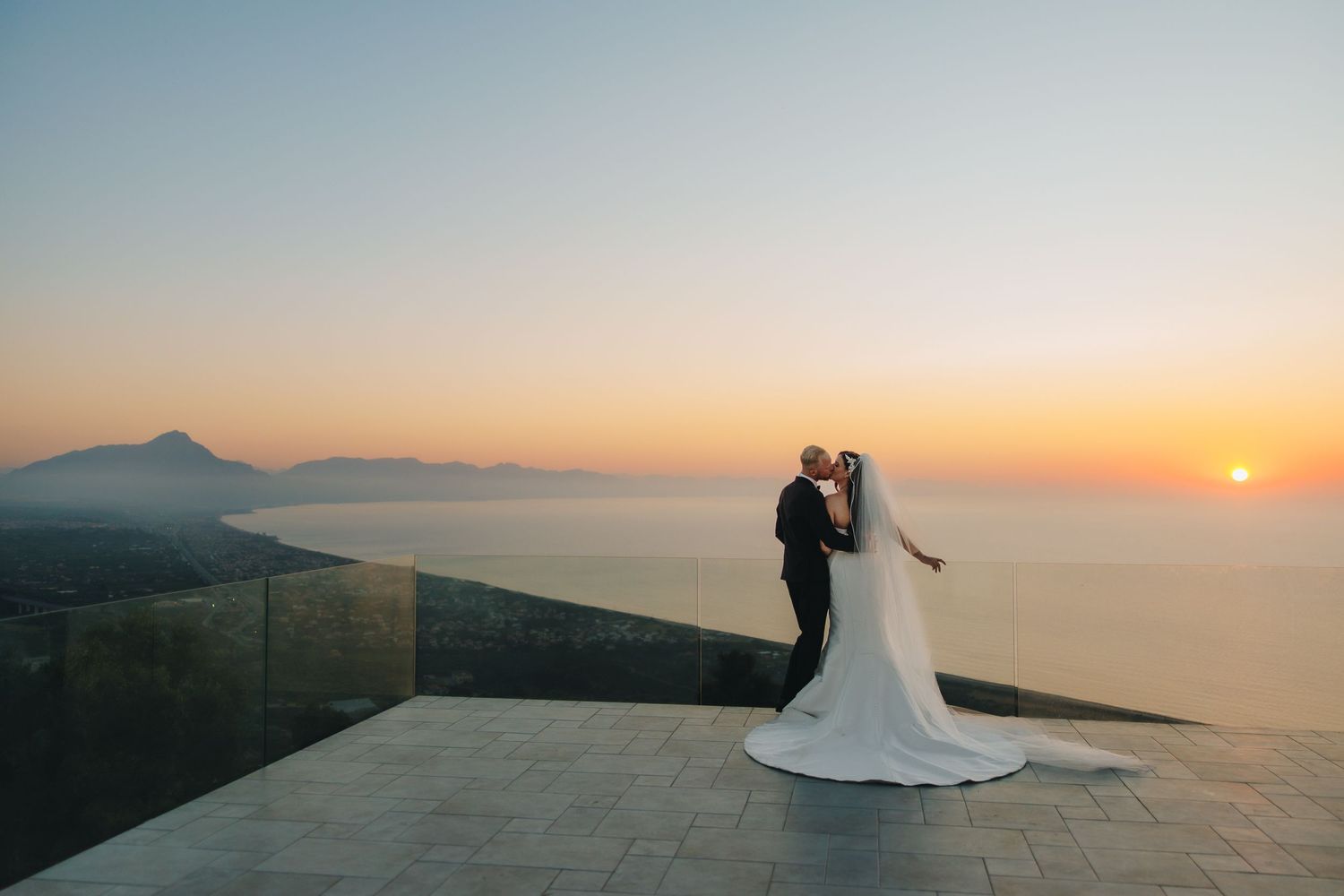 Bride and groom share a romantic kiss at sunset on a scenic overlook with mountain and ocean views.