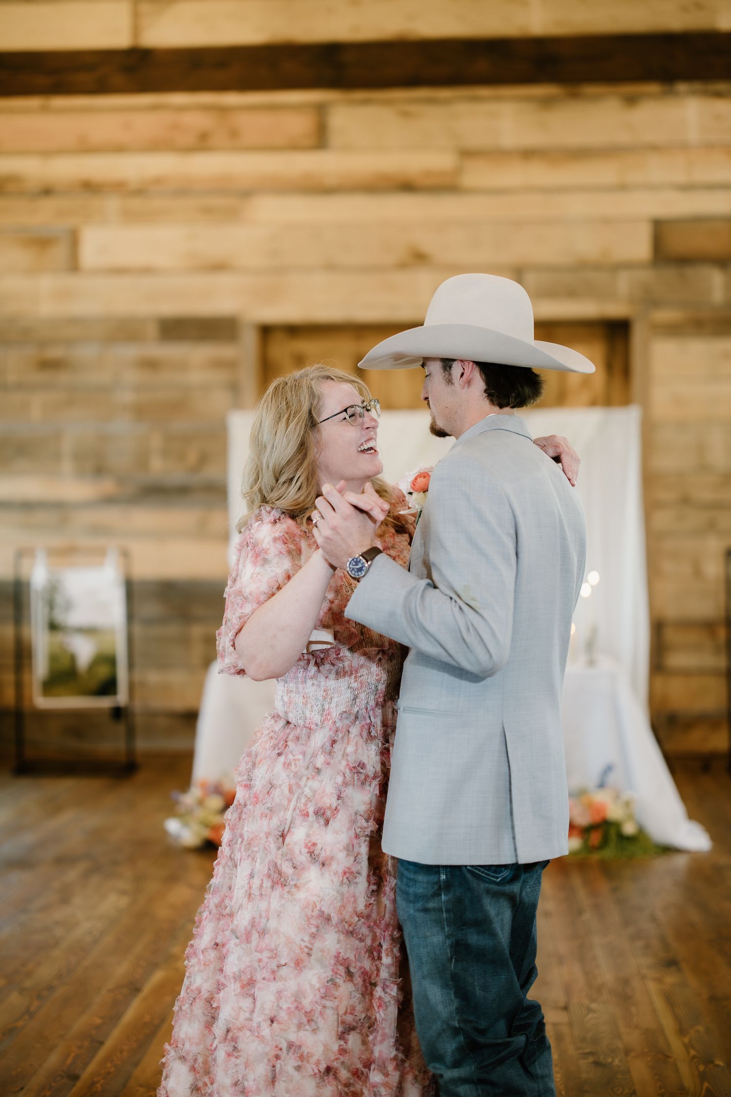 Groom in cowboy hat shares a mother-son dance inside a rustic wooden barn.