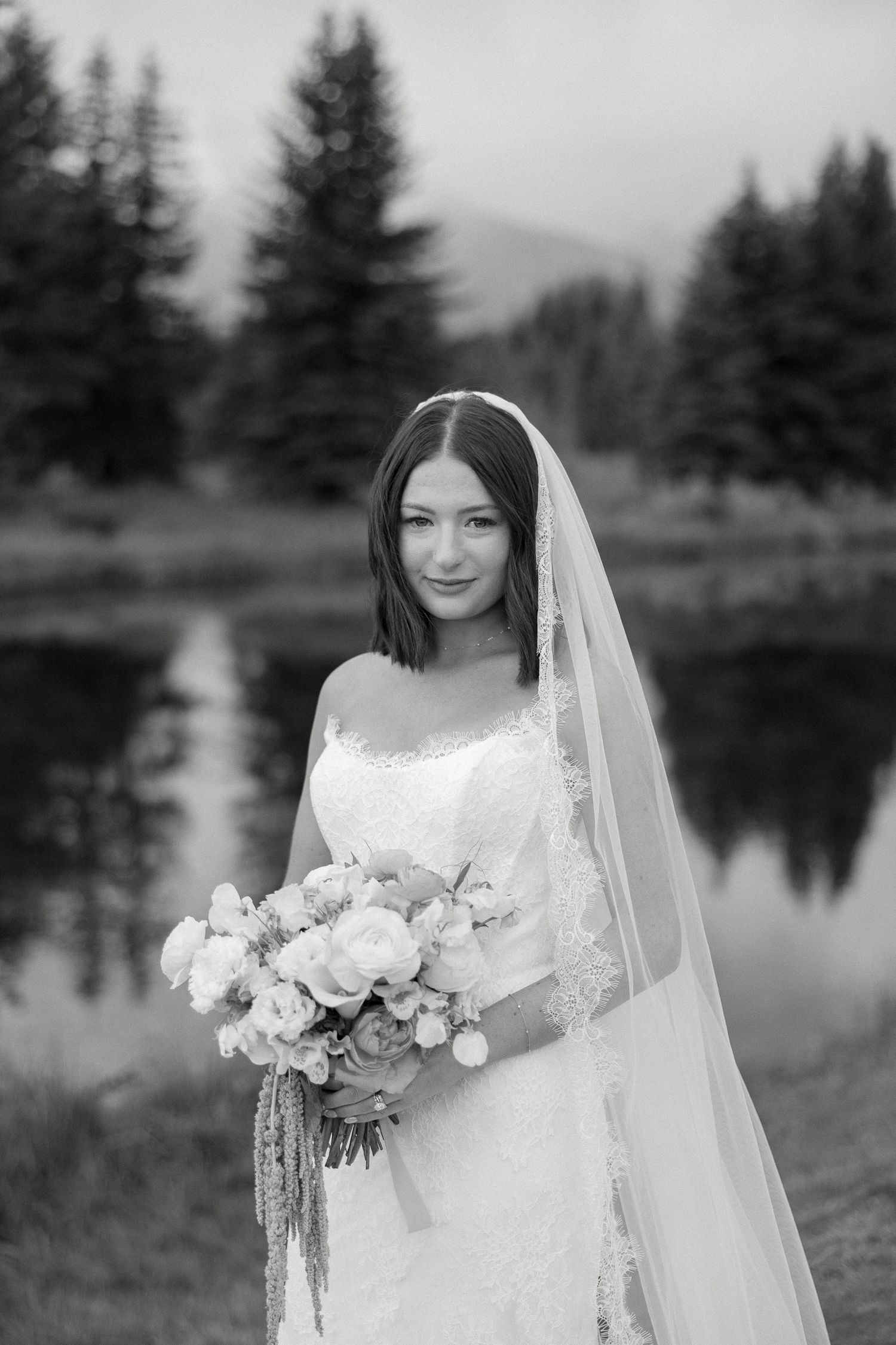 Black and white bridal portrait with veil and bouquet beside a reflective mountain lake.