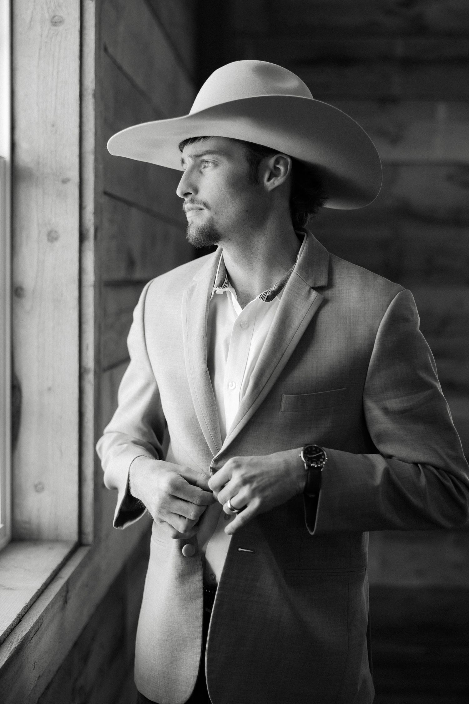 Stylish groom in gray suit and cowboy hat buttoning jacket by a window, black and white photo.