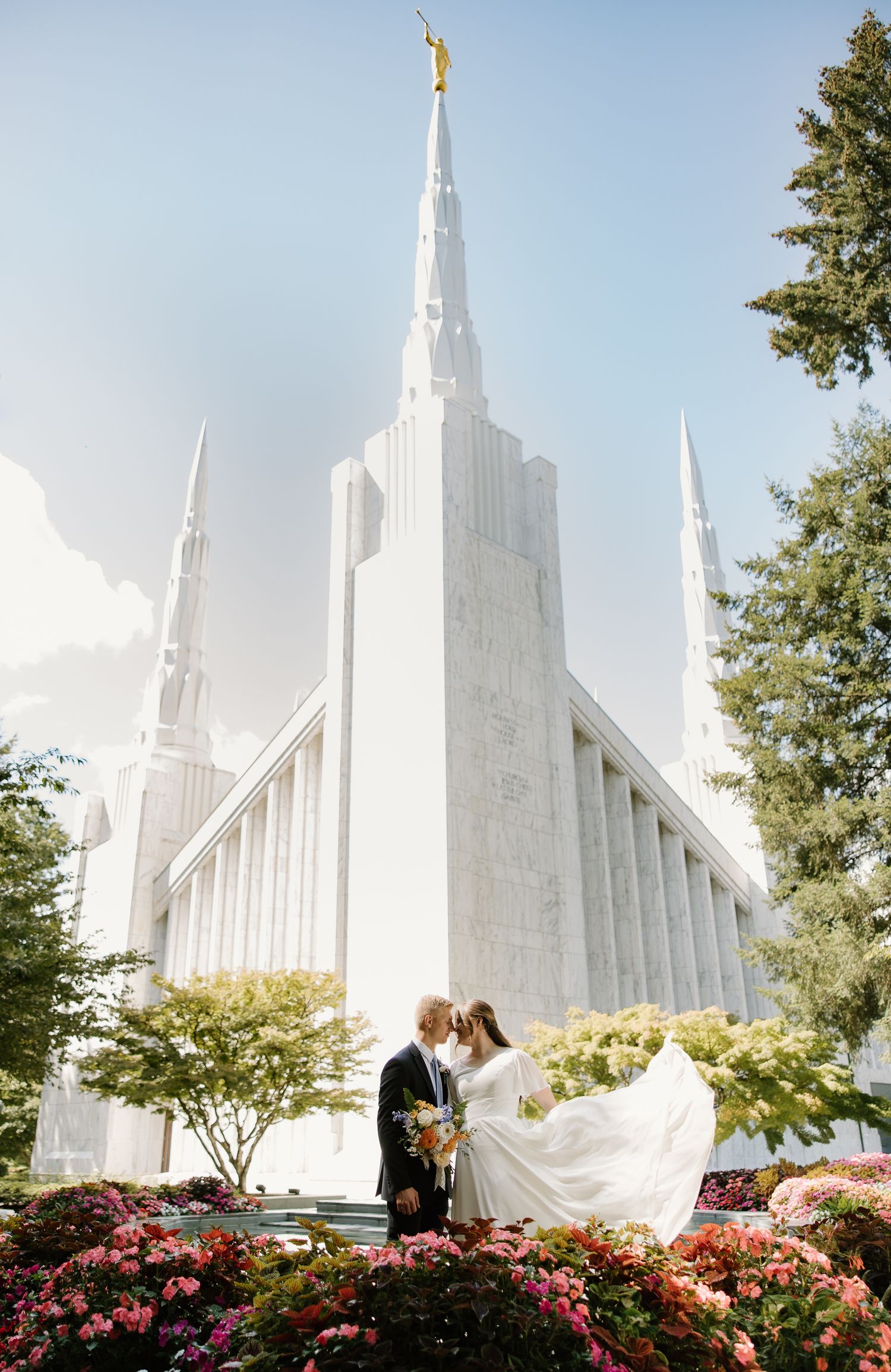 Bride and groom kiss in front of a stunning white LDS temple surrounded by colorful flowers.