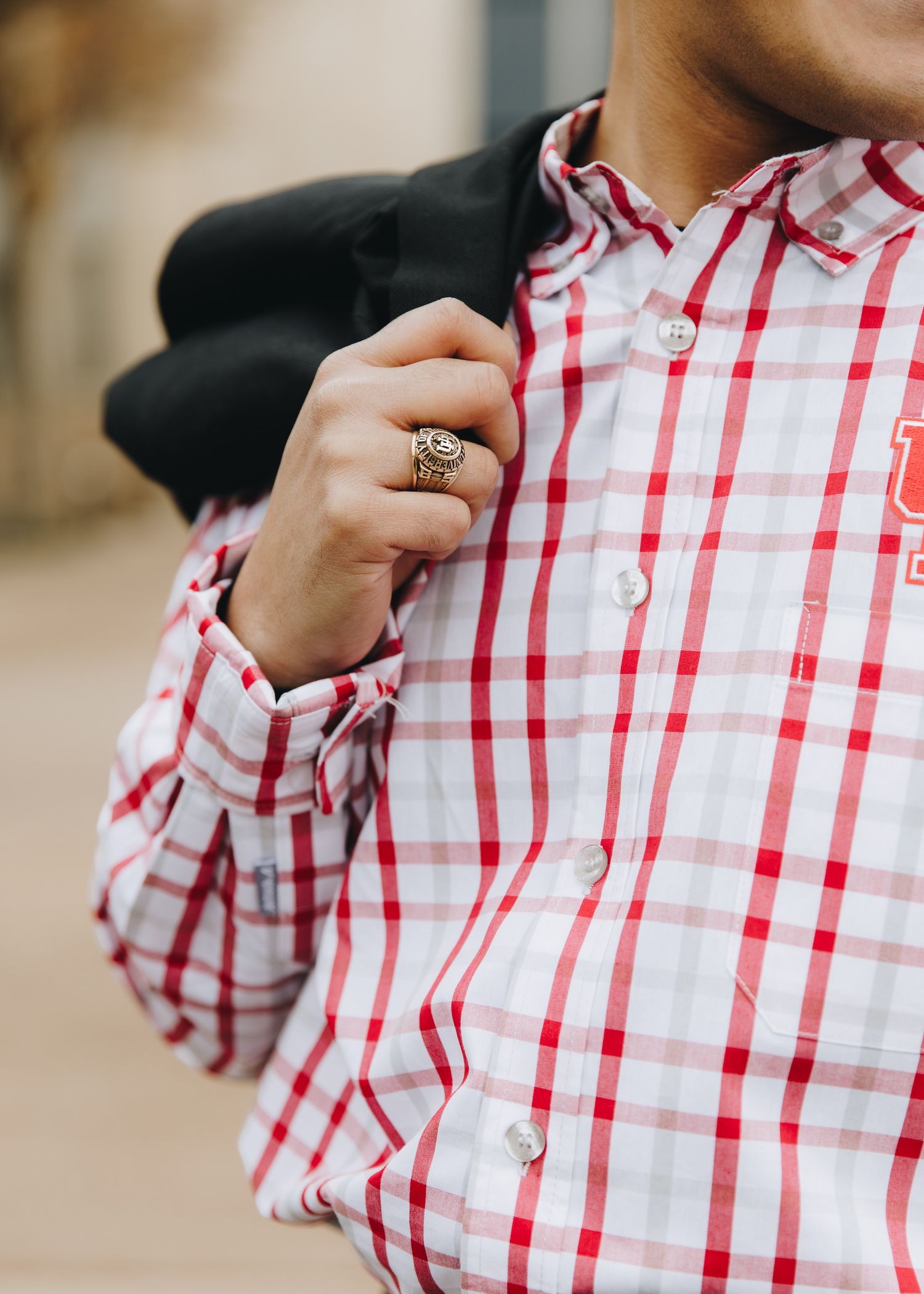 Close-up of man in red plaid shirt holding black blazer with gold class ring visible.