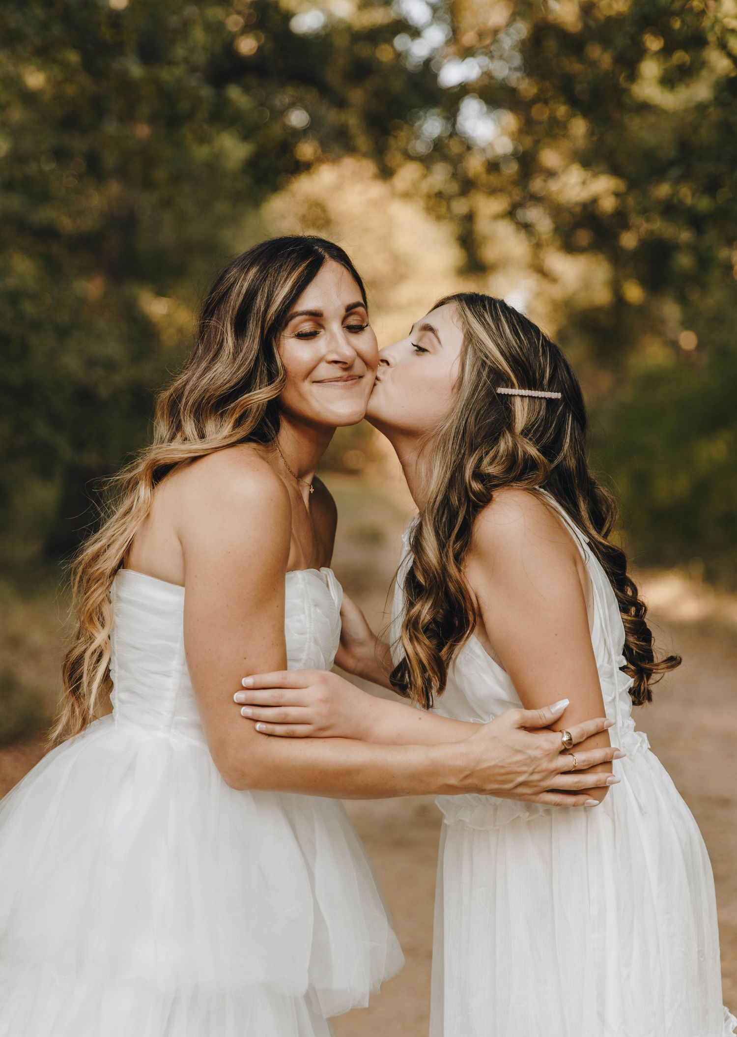 Two brides in white dresses share a tender kiss during an outdoor wedding ceremony.