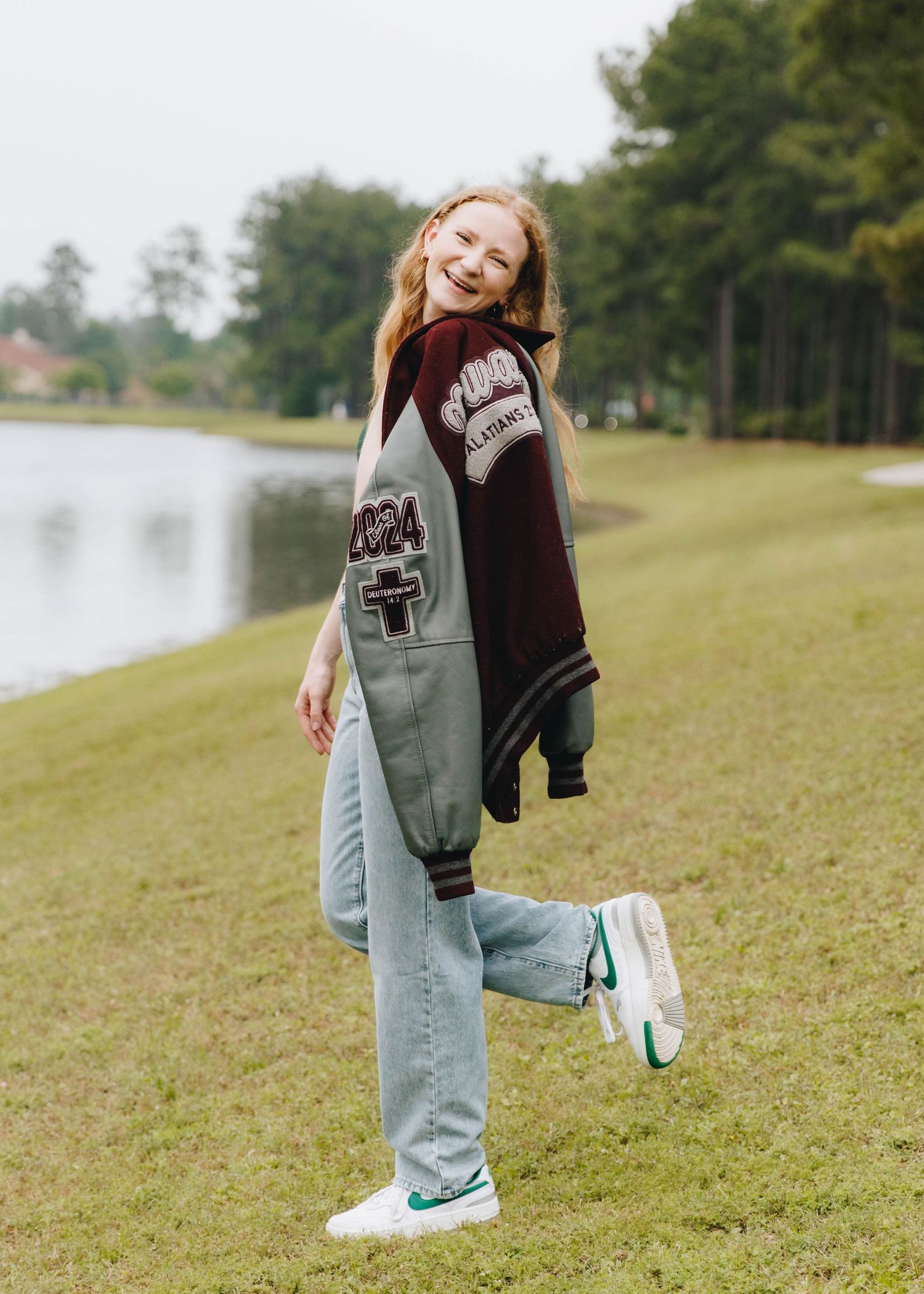 Smiling teen girl in letterman jacket and jeans posing playfully outdoors near a lake in senior photos.