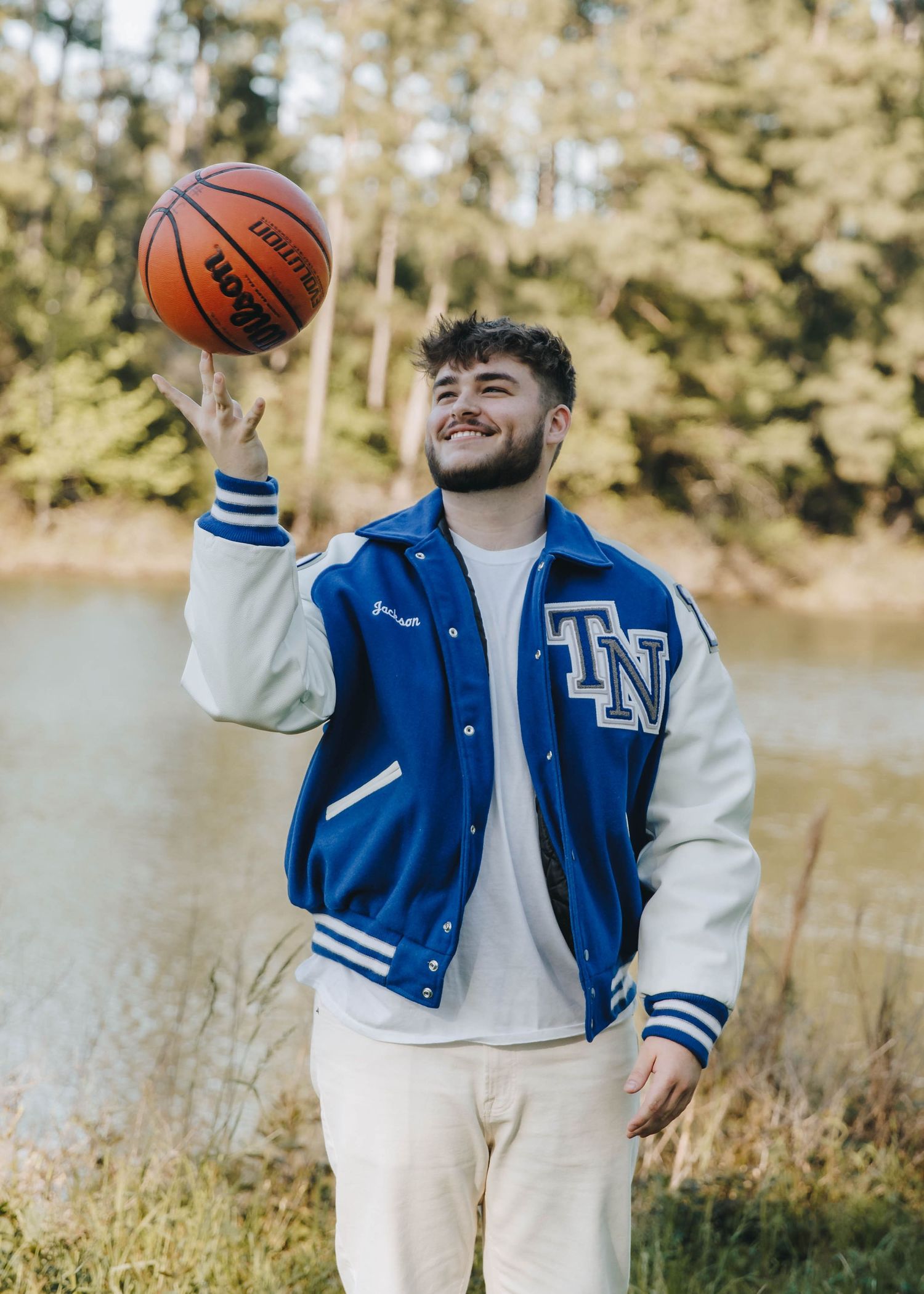Smiling young man spinning a basketball on his finger wearing a blue TN varsity jacket outdoors.