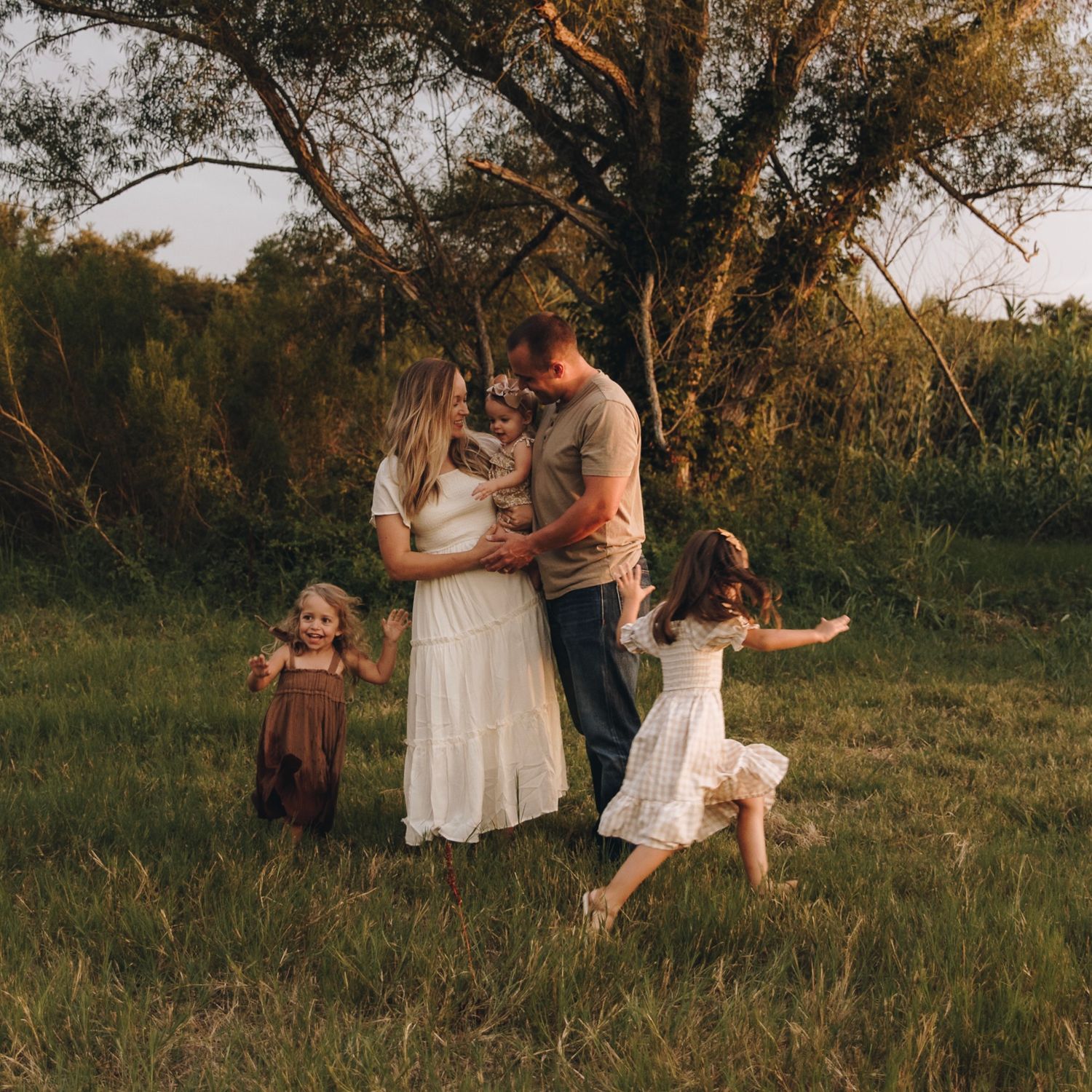 Candid family photo of parents with baby and two daughters playing in sunlit green meadow at dusk.
