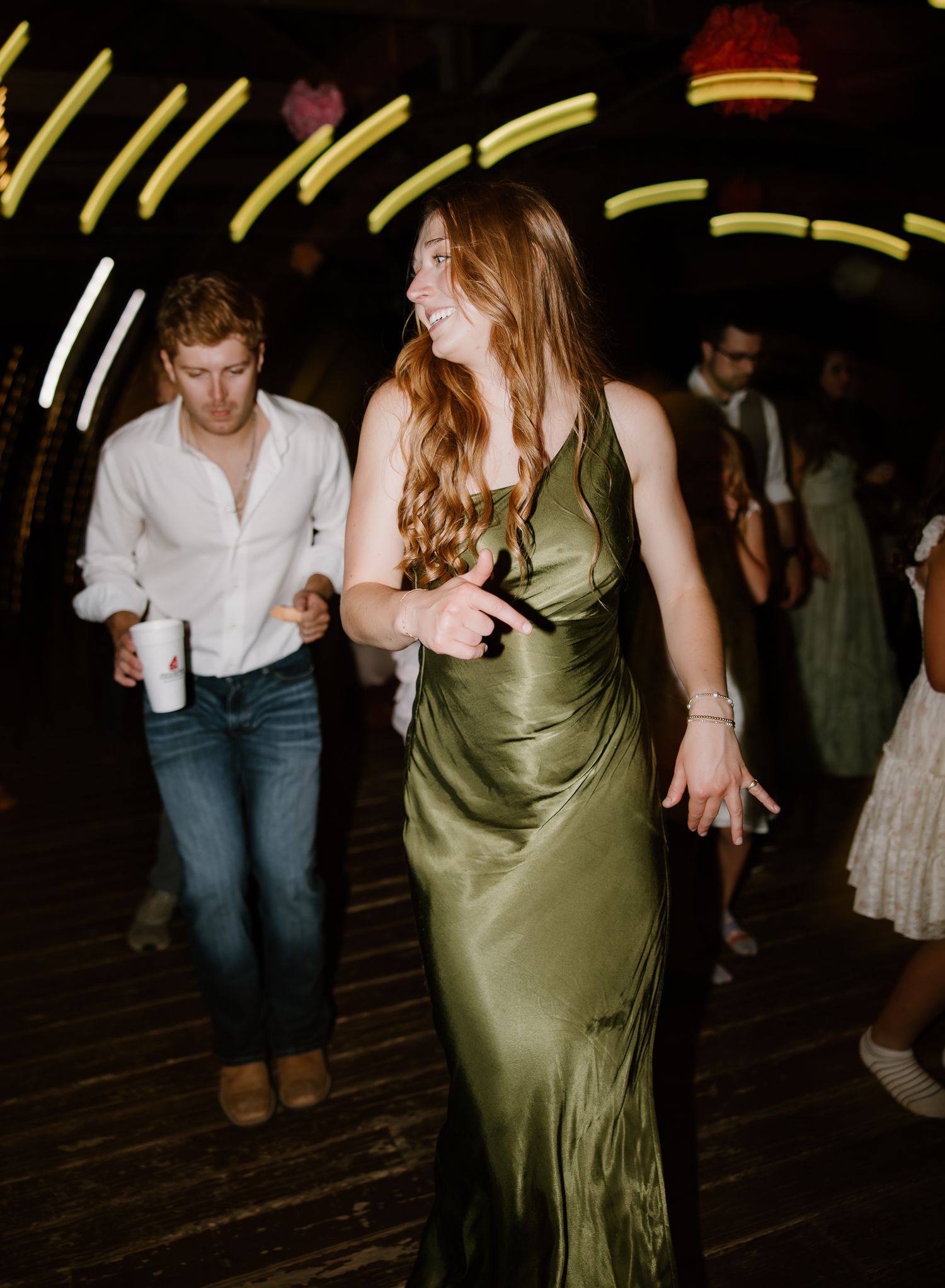Woman in elegant olive green satin gown dancing joyfully at lively wedding reception with neon lights.