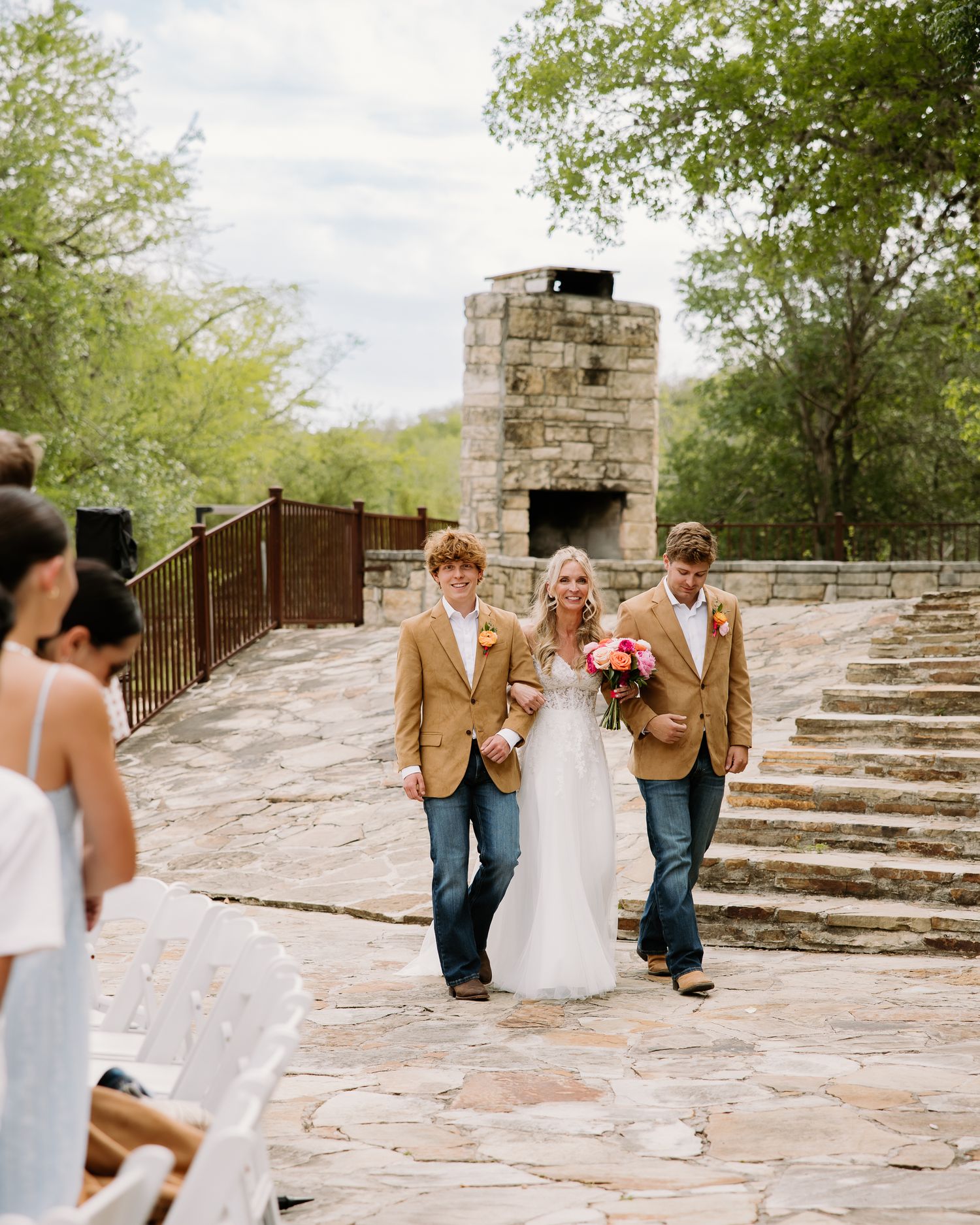 Bride walks down stone aisle escorted by two young men in tan blazers and jeans at outdoor wedding.
