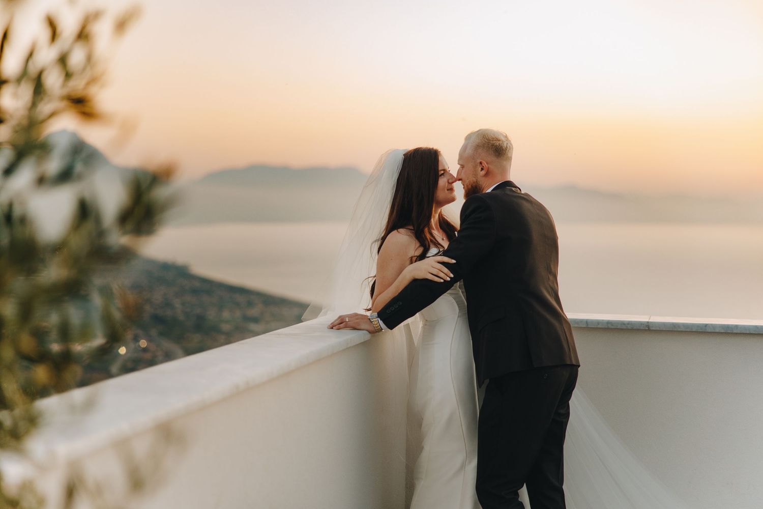 Couple sharing a romantic kiss on a white balcony overlooking the sea at sunset in Santorini.