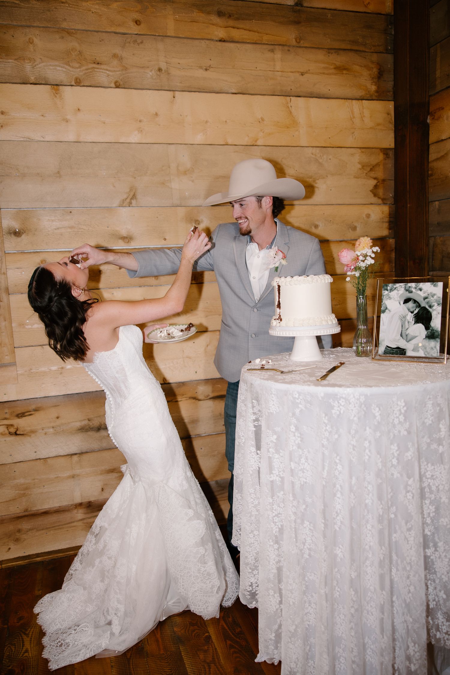 Bride and groom playfully feed each other wedding cake in a rustic barn reception venue.