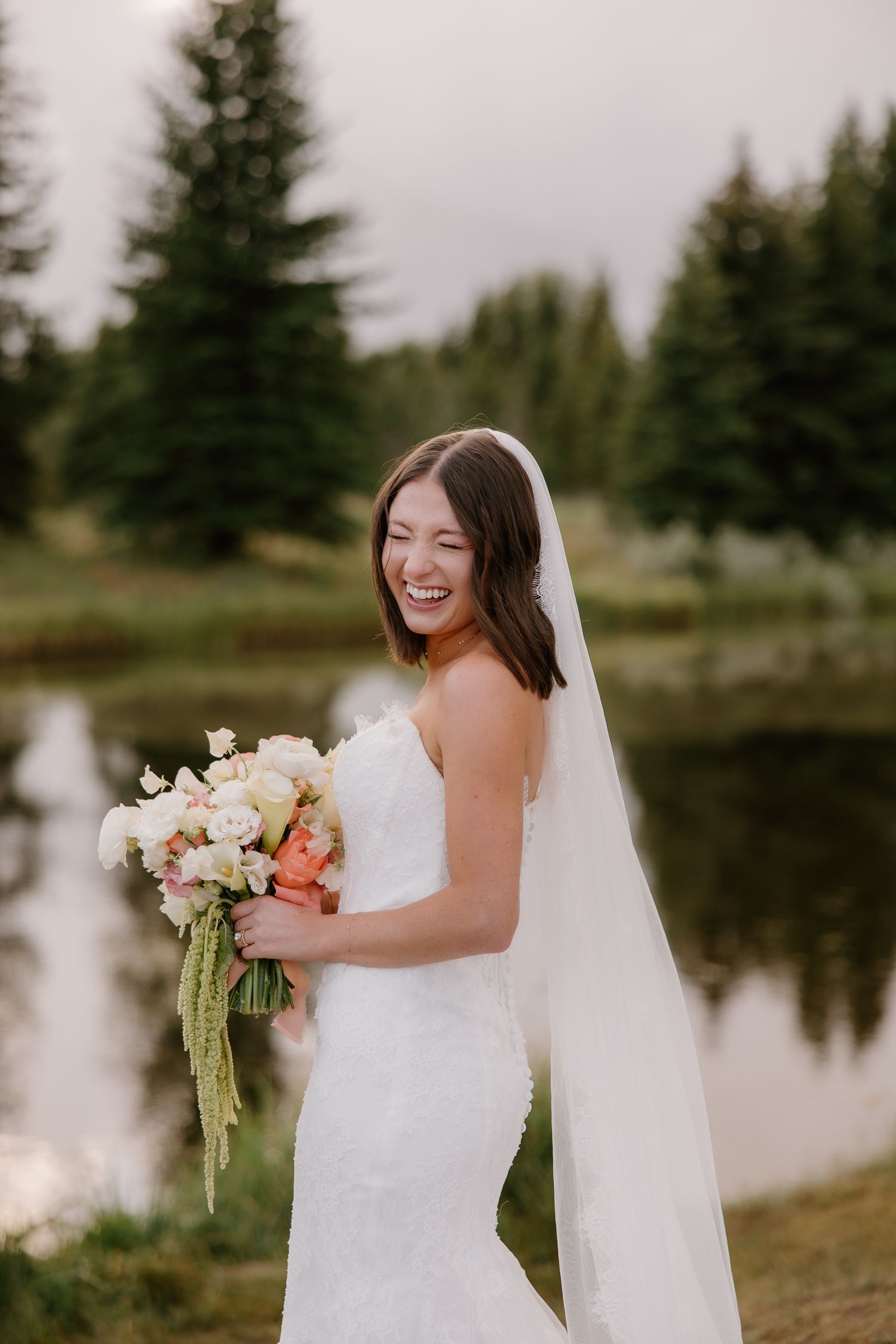 Laughing bride in strapless lace gown holding peach and white bouquet by serene forest pond.
