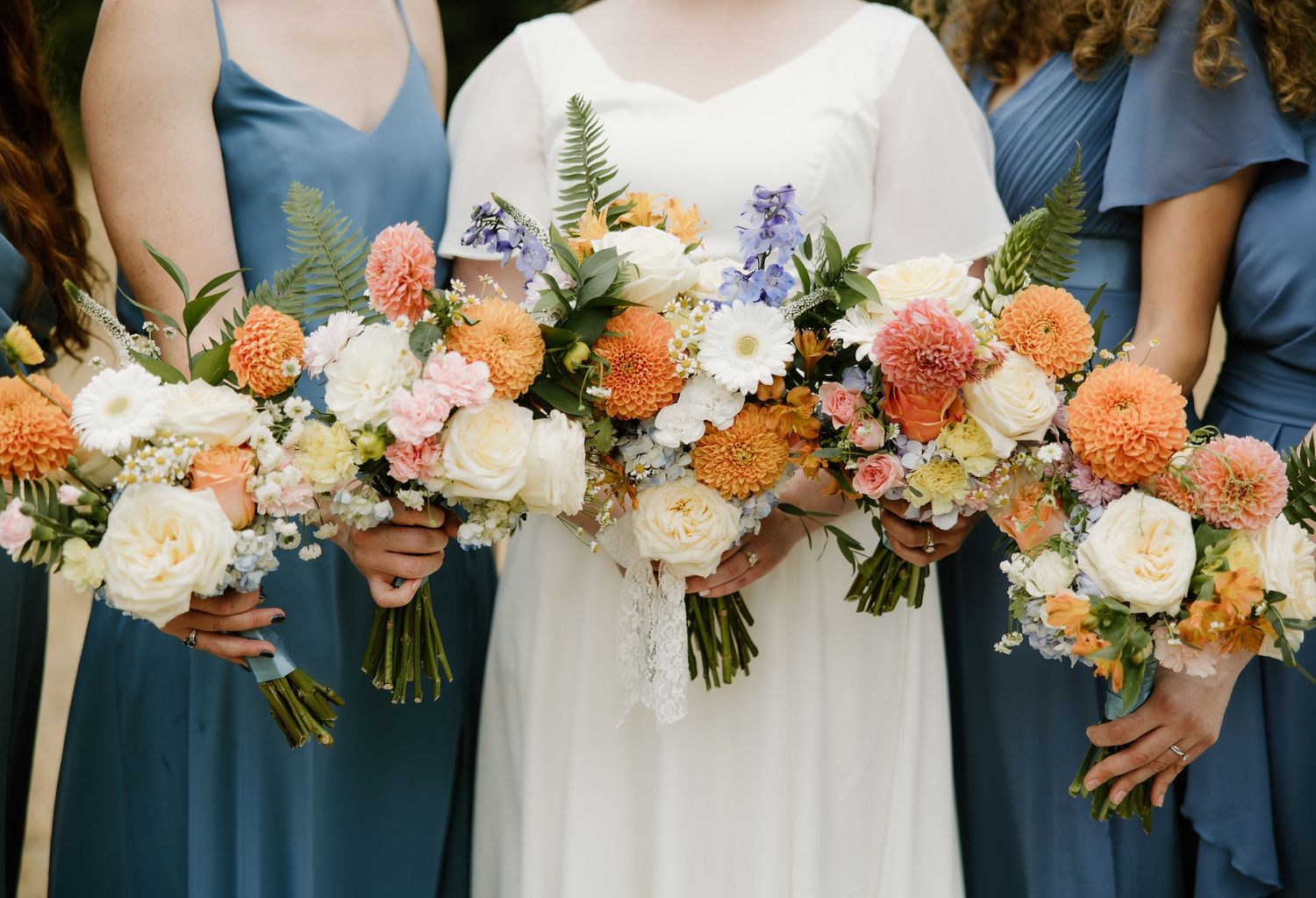 Bride and bridesmaids in dusty blue dresses display stunning orange, white, and blue floral bouquets.