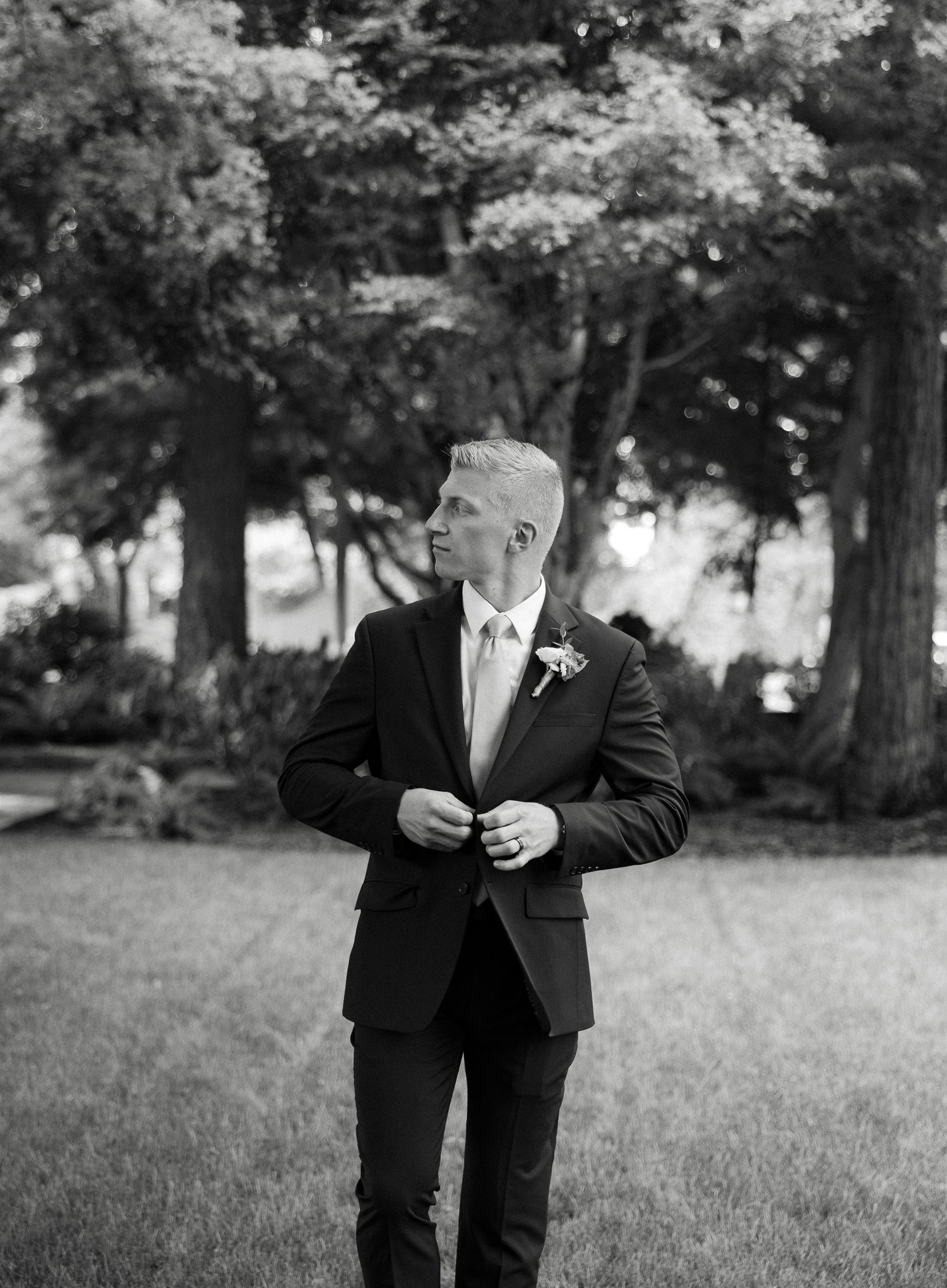 Elegant black and white portrait of a groom in a suit adjusting his jacket outdoors.