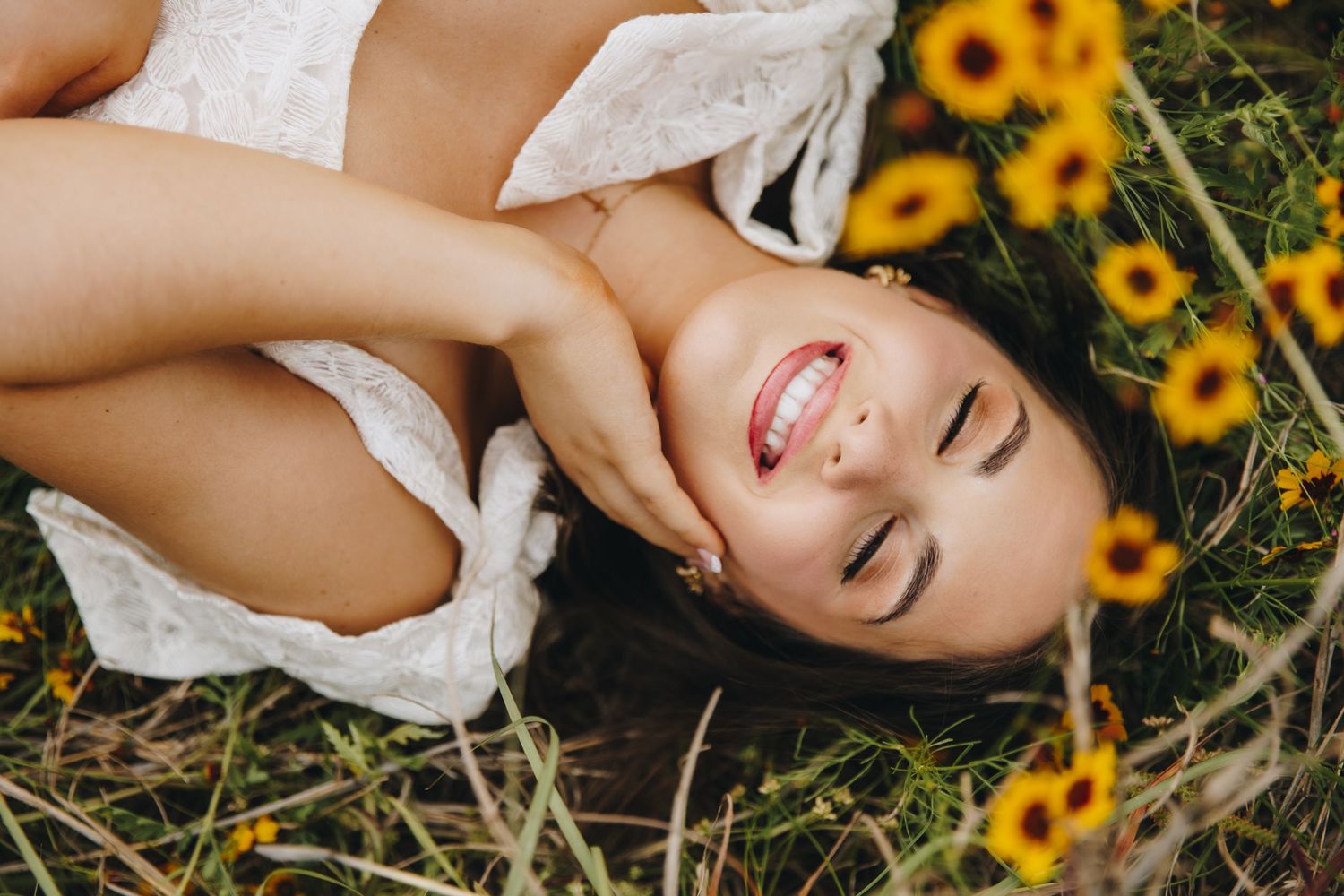 Smiling woman in white dress lies in sunflower field laughing with eyes closed.
