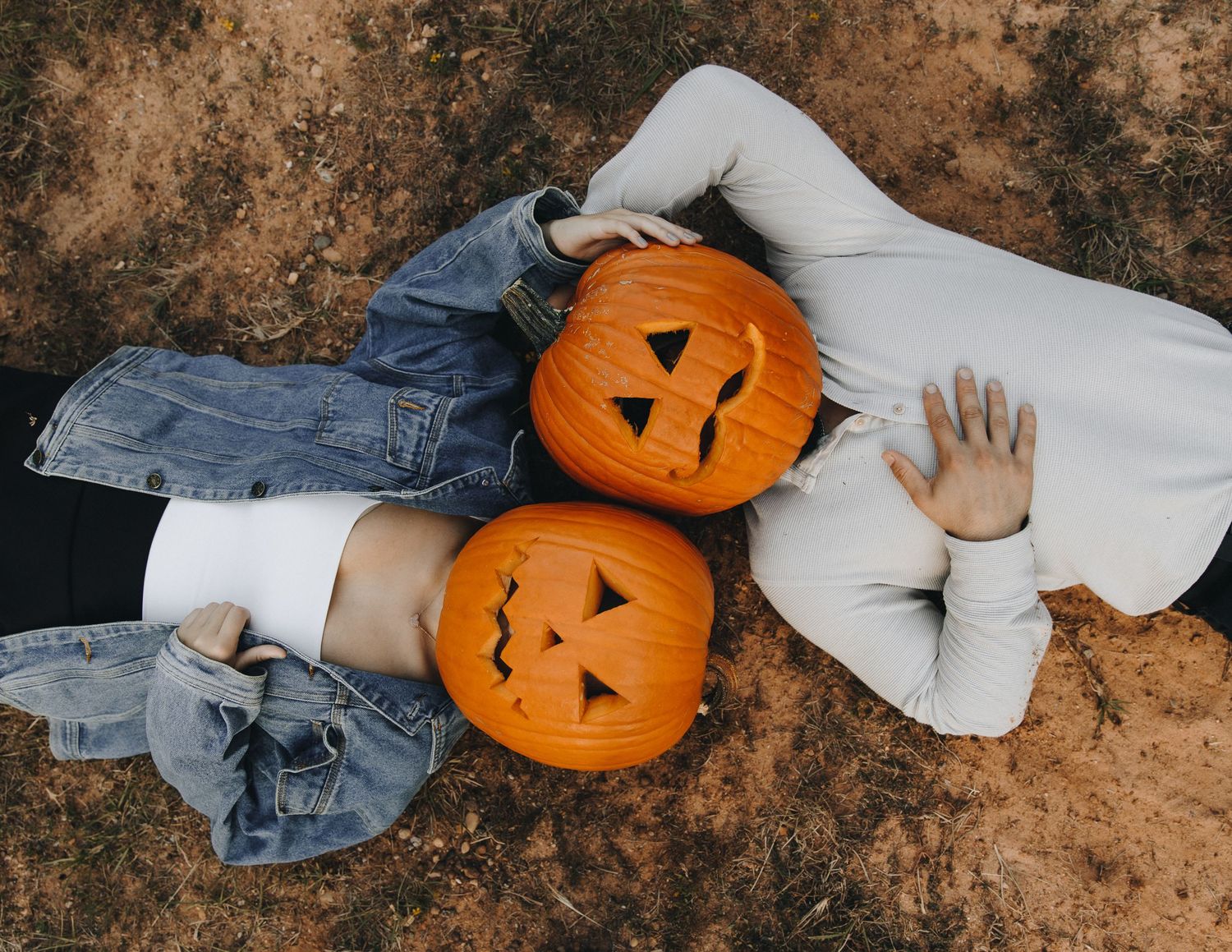 Two children lay on the ground with carved Halloween jack-o-lantern pumpkins covering their faces outdoors.