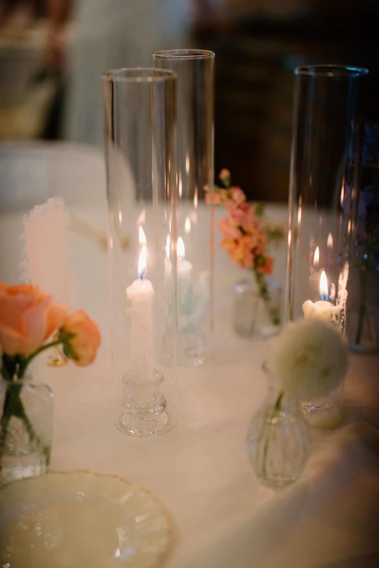 Elegant wedding table centerpiece with lit taper candles in glass holders and pink roses on white linen.