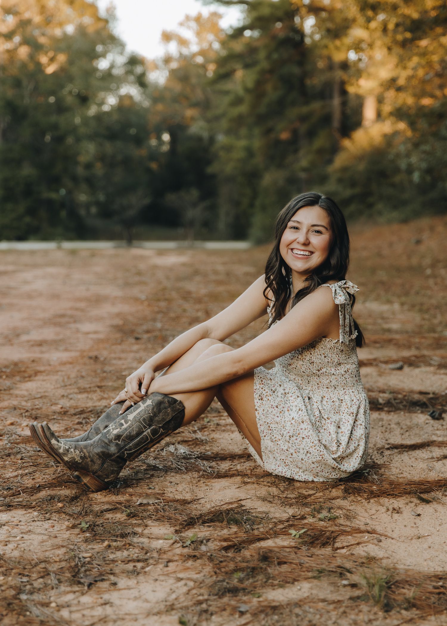 Smiling young woman in floral dress and cowboy boots sitting on a dirt path in golden hour light.