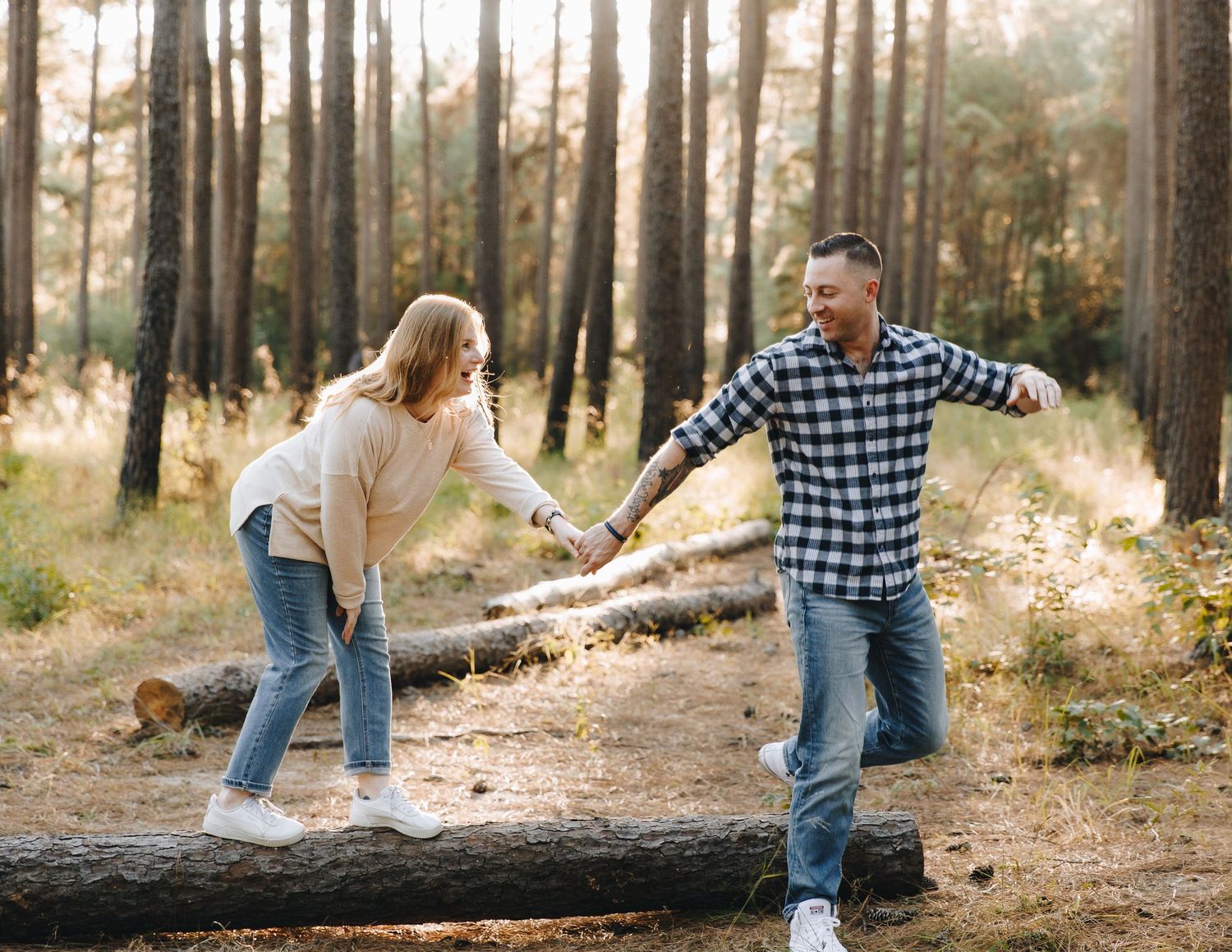 A cheerful couple holds hands and balances on a fallen log in a sunlit pine forest.