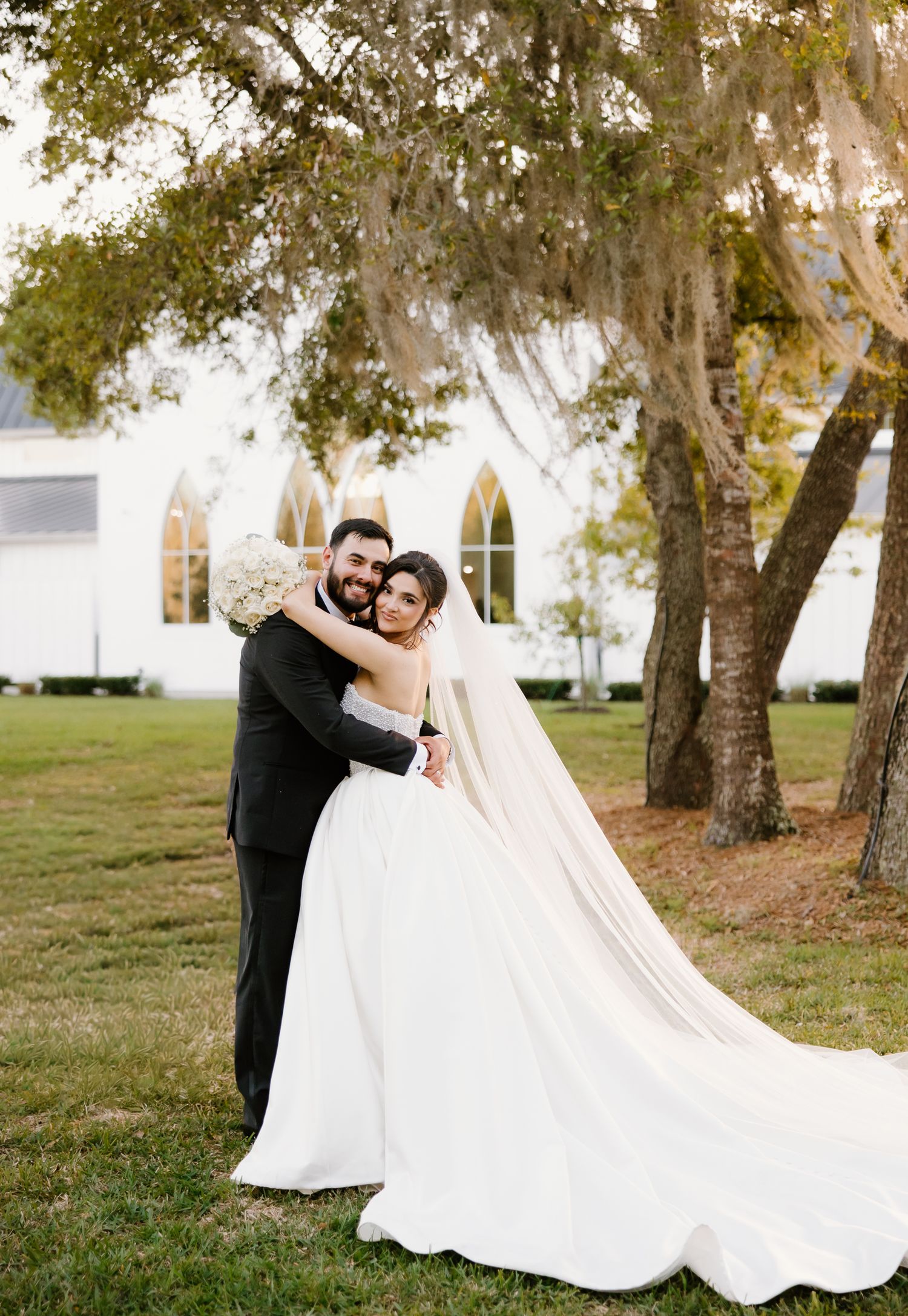 Bride and groom embrace on lawn before a white chapel with gothic arched windows.