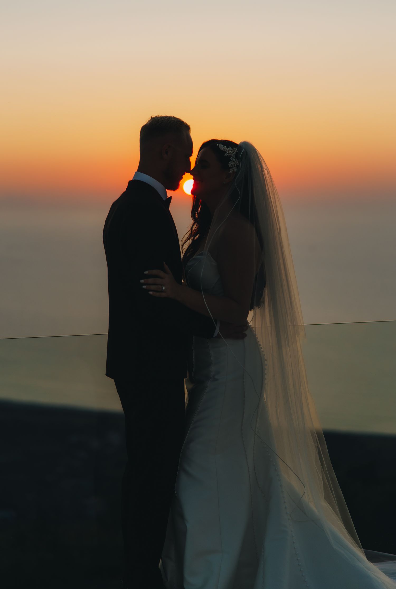 Romantic silhouette of bride and groom sharing a kiss against a vibrant ocean sunset.