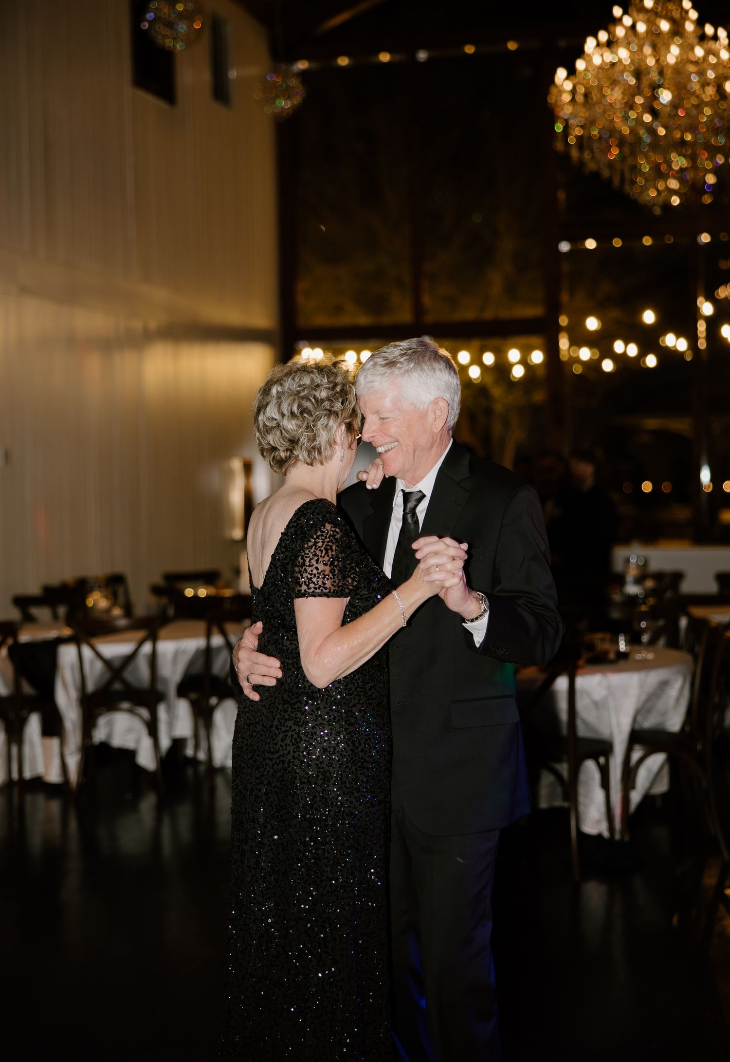 Elegant couple dancing at a formal wedding reception with chandeliers and string lights.