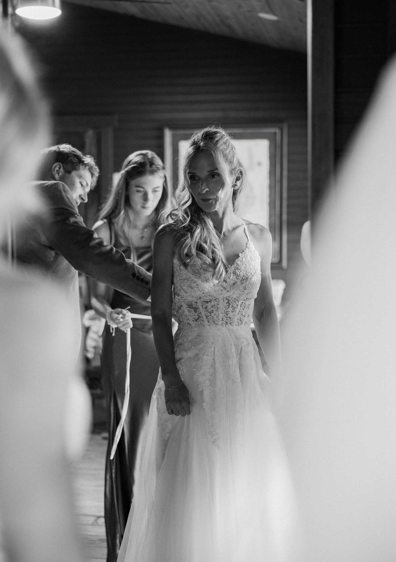 Bride smiling in lace wedding gown while bridesmaids help her dress in black and white photo.