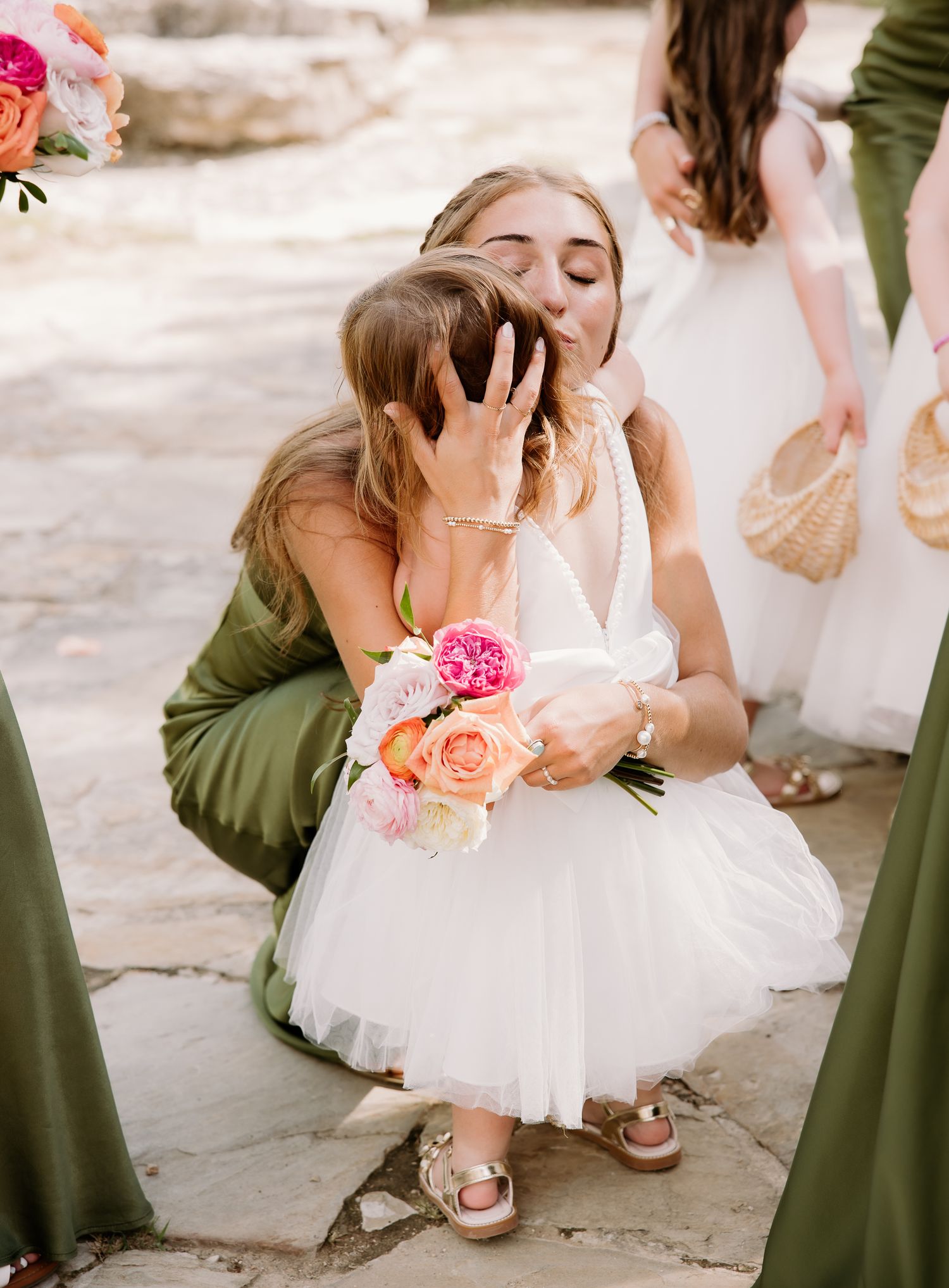 Bride in green dress kneeling to kiss flower girl in white dress holding colorful pink and peach bouquet outdoors.