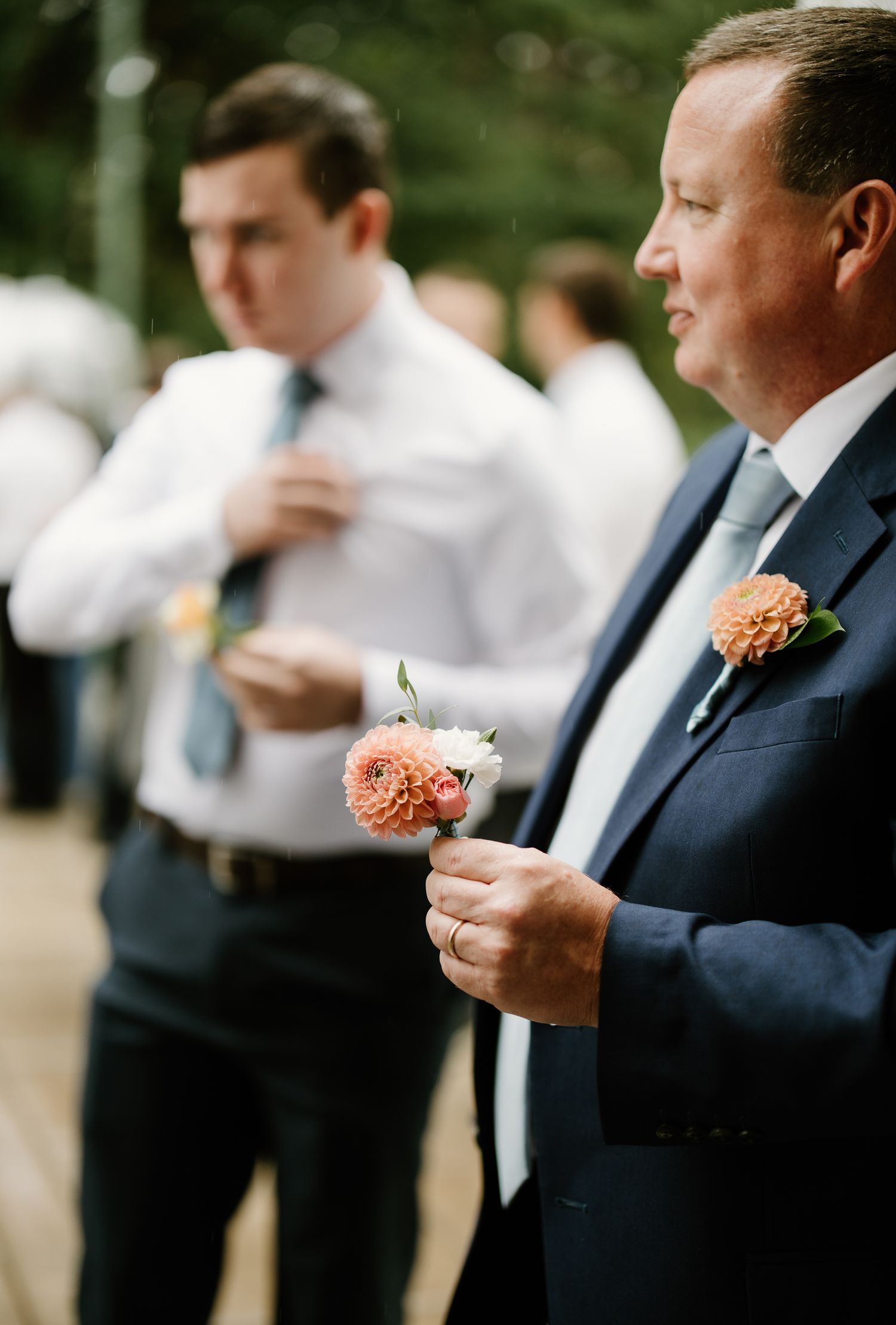 Man in navy suit holds coral boutonniere at wedding, with groomsman adjusting tie in background.