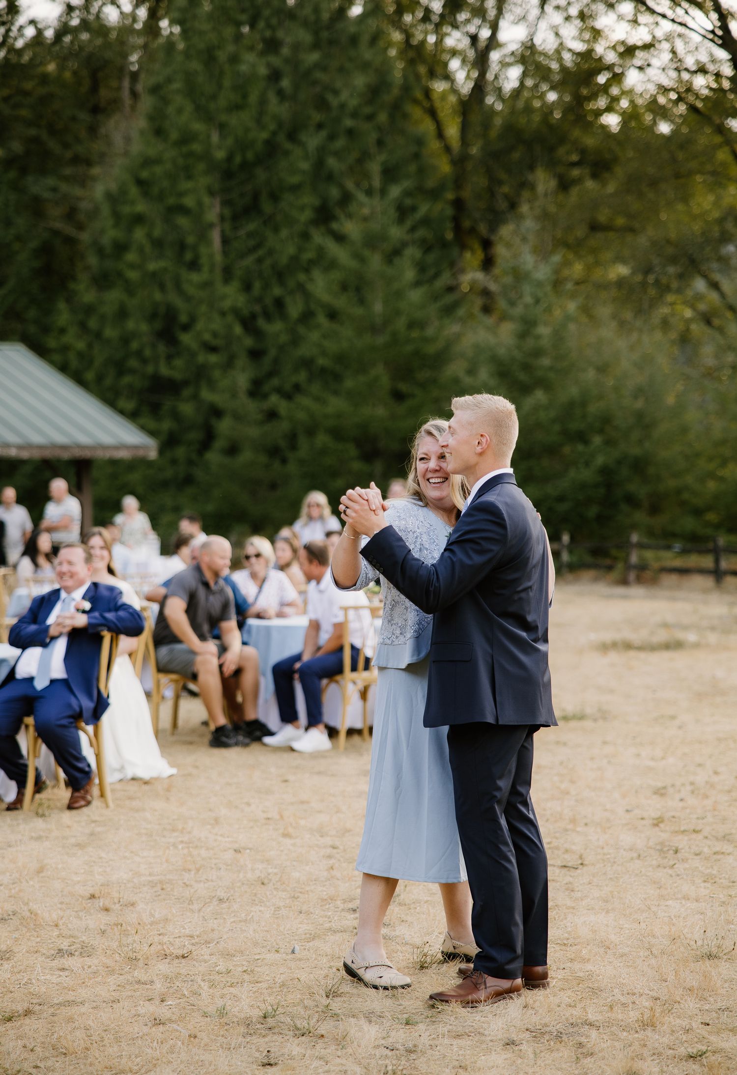 Groom dances with mother in light blue dress at outdoor wedding reception with seated guests in background.