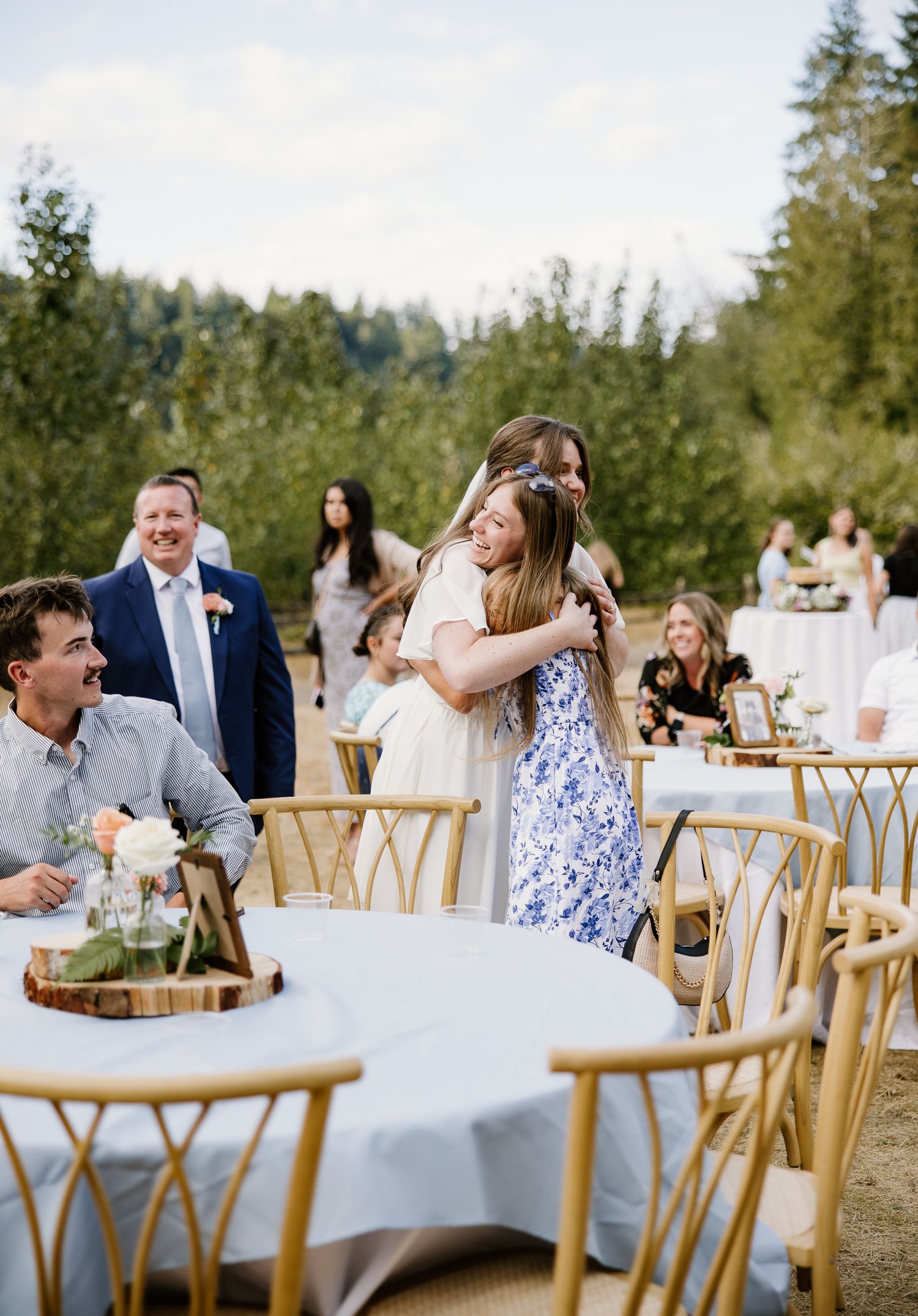 Bride in white dress hugs guest at outdoor wedding reception with gold chiavari chairs.
