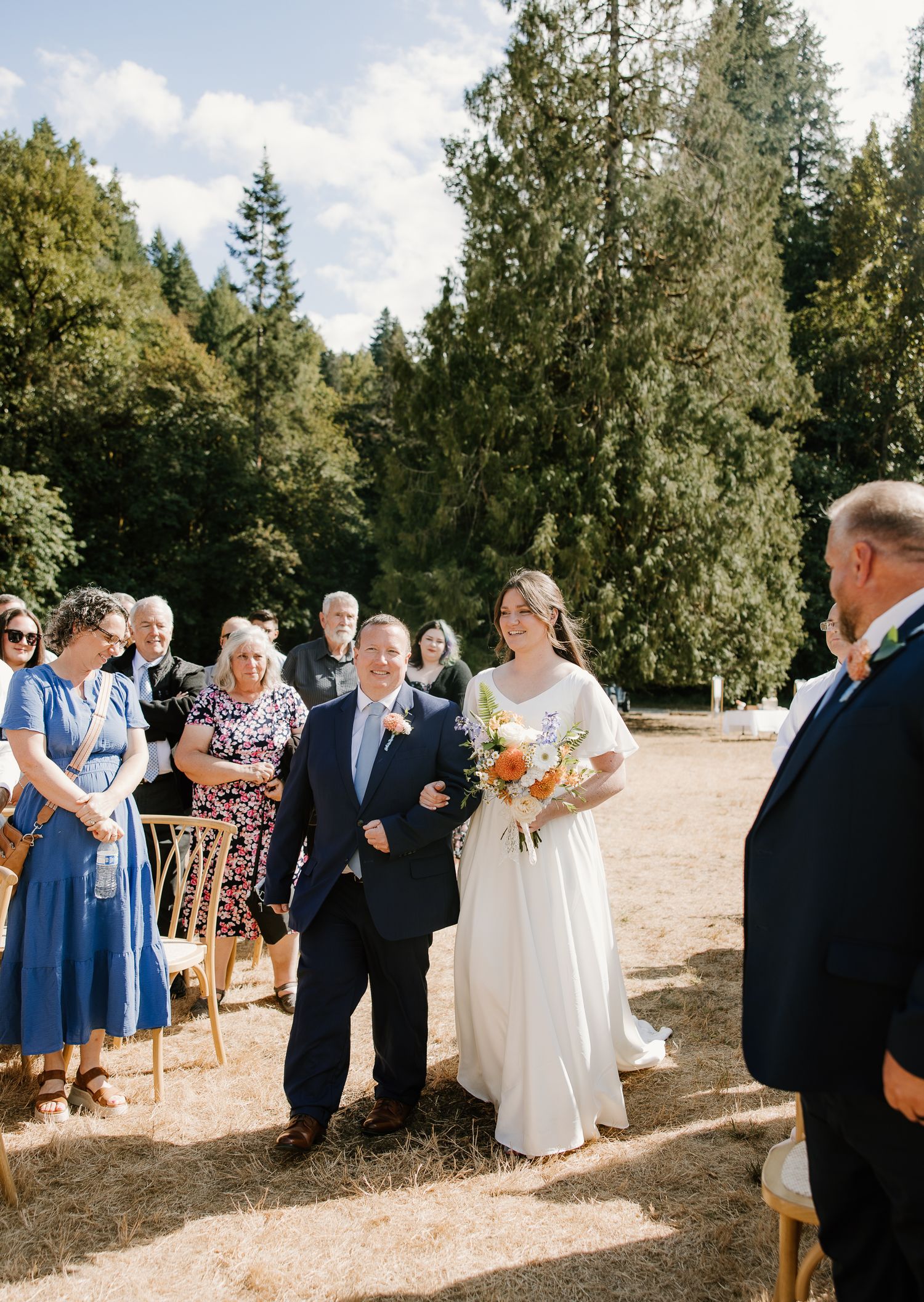 Bride walks down outdoor aisle with escort during sunny forest wedding ceremony.