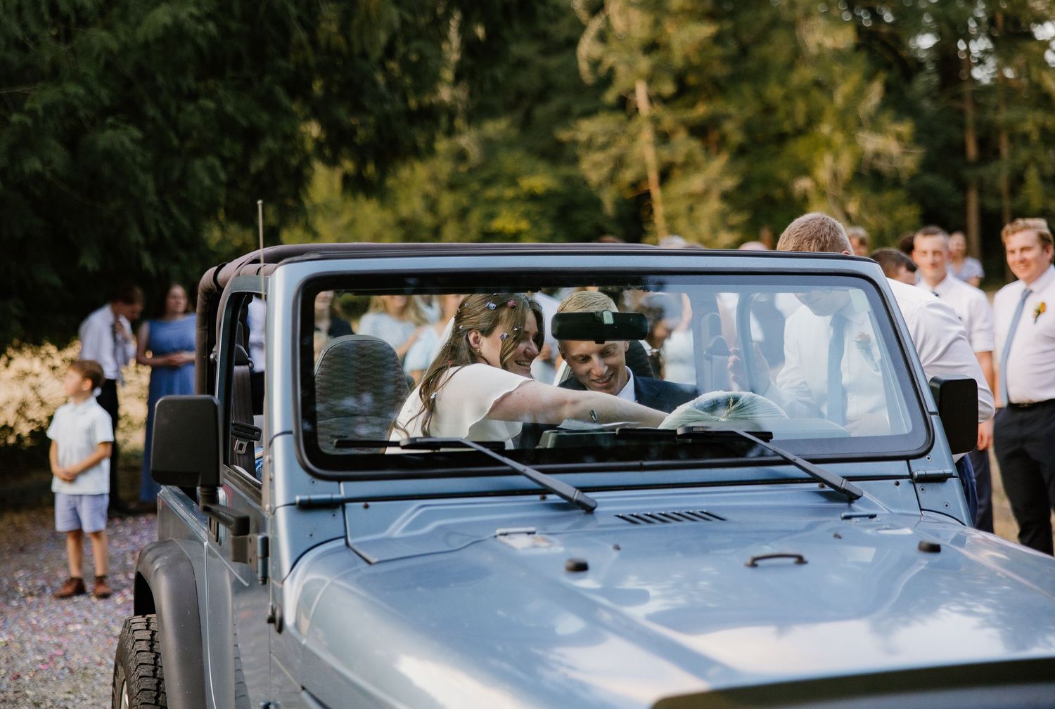 Bride and groom sit together in a gray open-top Jeep Wrangler surrounded by wedding guests outdoors.