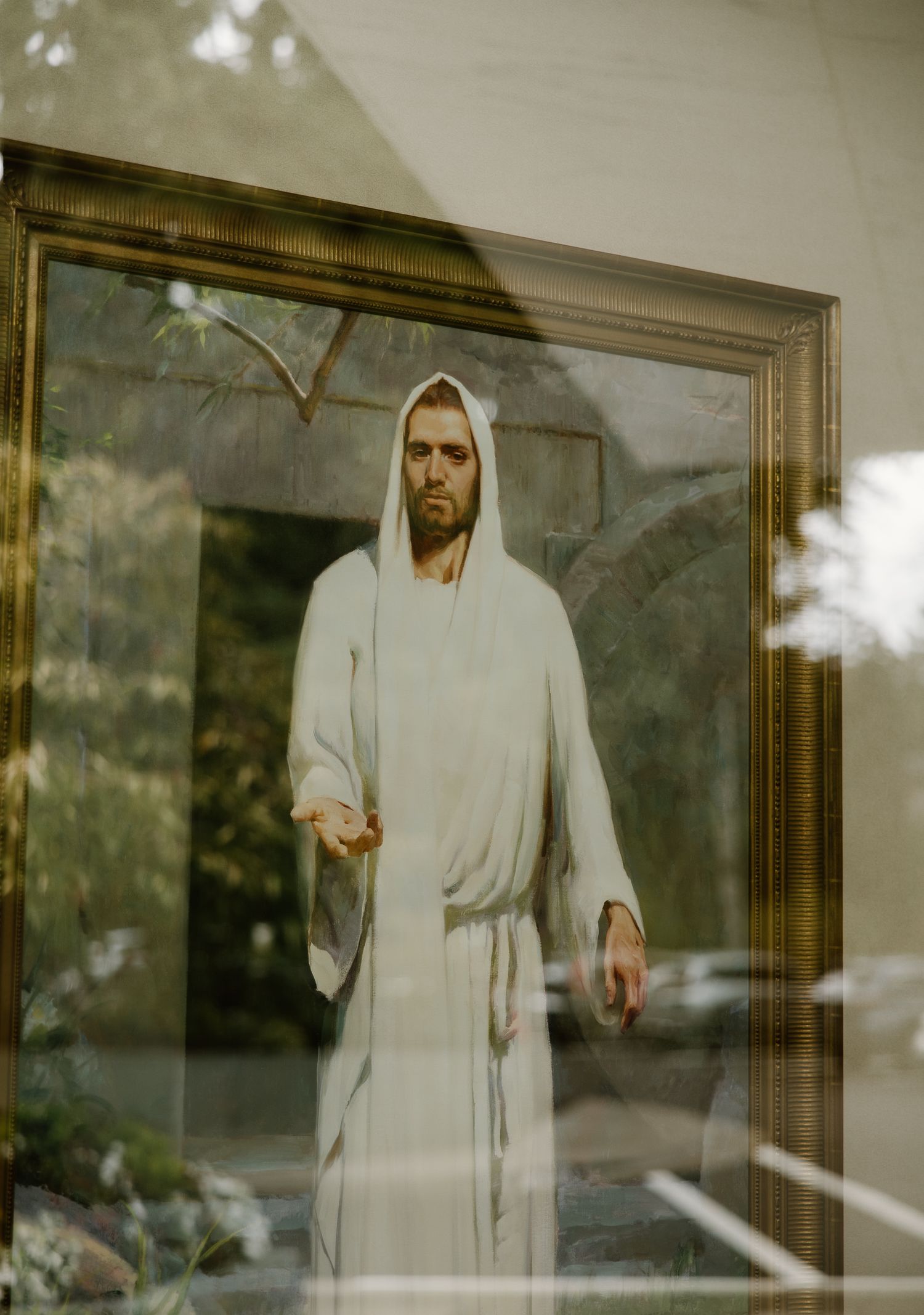 Framed painting of Jesus Christ in white robes displayed behind glass with outdoor reflections.