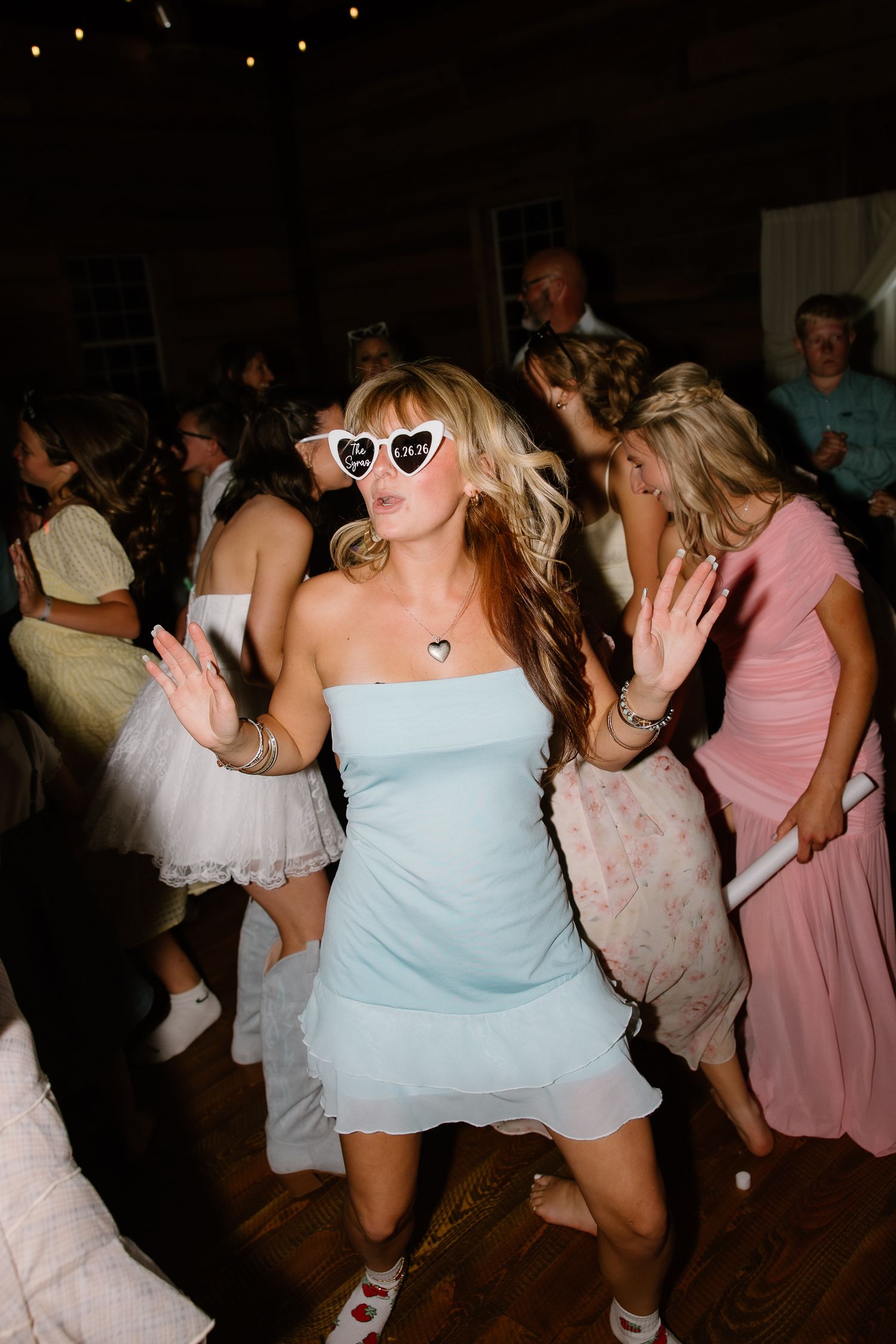 Guest in a light blue dress and heart-shaped sunglasses dancing at a lively barn wedding reception.