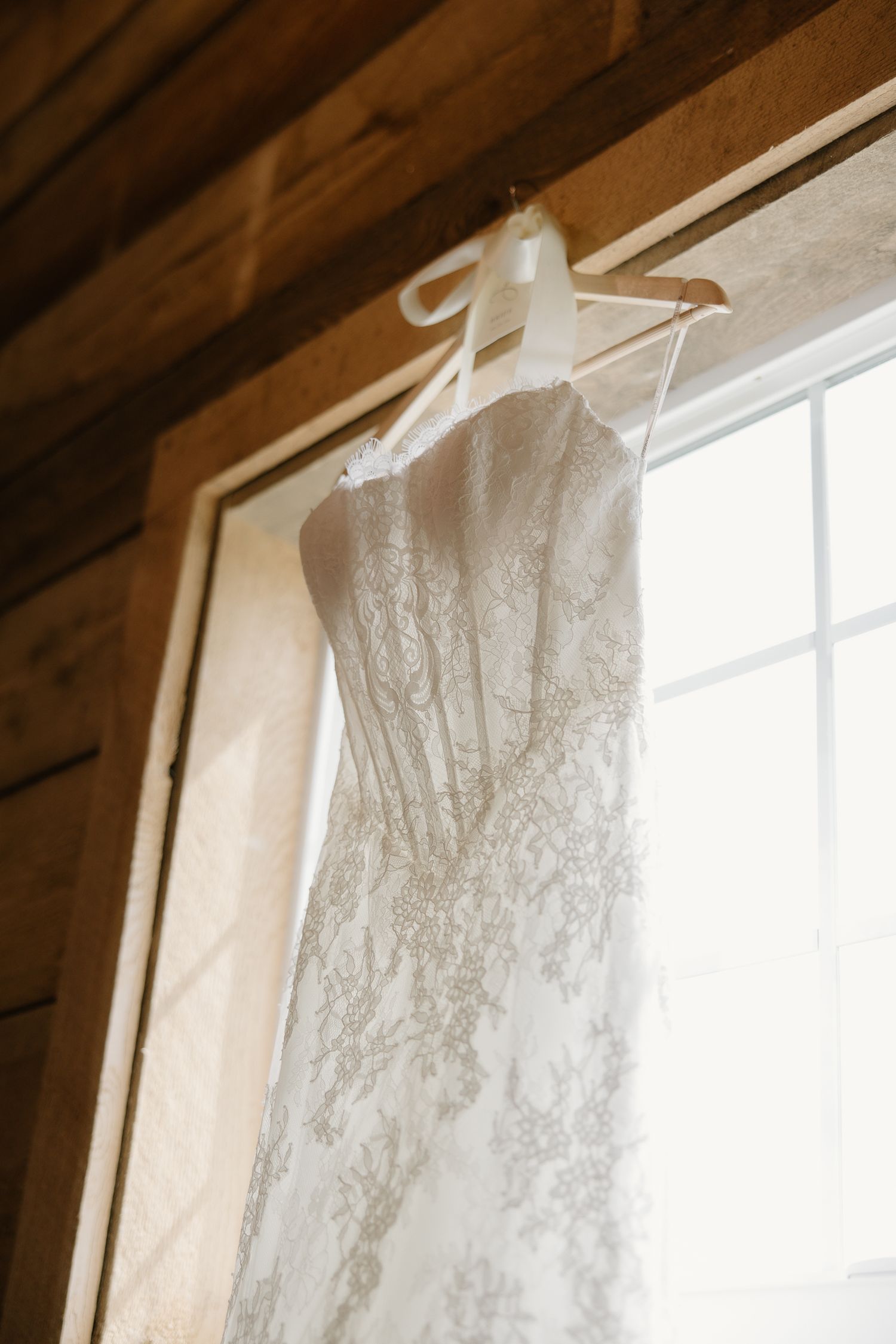 Elegant lace wedding dress hanging on a white hanger in front of a sunlit window in a rustic wooden cabin.